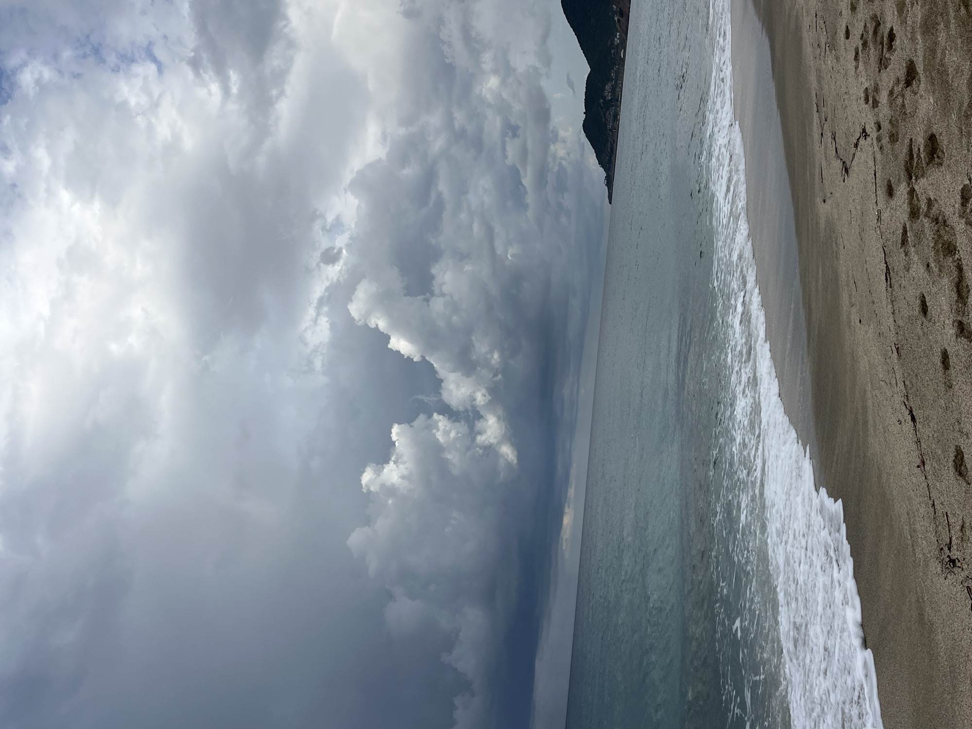 The image shows a beach scene in Alanya, Turkey. The sky is filled with dark, stormy clouds. A body of water borders the sandy beach, and waves are crashing onto the shore. A dark, mountainous landmass is visible in the distance. Footprints are visible in the sand. There are no visible people in the picture.