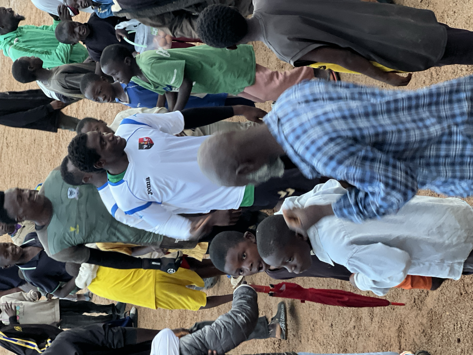 The image depicts a group of people, mostly children, gathered outdoors on a dusty surface, likely a sports field or undeveloped area. The environment is arid and appears to be in Jos, Nigeria, indicated by the local clothing styles and the overall scene.
In the foreground, a person wearing a blue and white checkered shirt is turned away from the camera, their back to the viewer. To their left, a young boy in a light-colored shirt looks up, appearing surprised. Adjacent to him, another child is partially visible, wearing a dark shirt. A red umbrella is held vertically by someone unseen in the lower left portion of the frame.
Dominating the center of the image is a man wearing a white and blue soccer jersey with the logo "Joma" visible. He is smiling and interacting with the people around him. To his left, a man in a green jersey is also visible. Several other individuals, primarily young boys and girls, are scattered throughout the scene, some looking towards the man in the soccer jersey, others engaged in their own interactions or simply observing.
The ground is covered with loose dirt and sand. The lighting suggests an outdoor setting, likely during daylight hours. The overall atmosphere appears to be one of community gathering and possibly a sports-related event, given the presence of soccer attire.