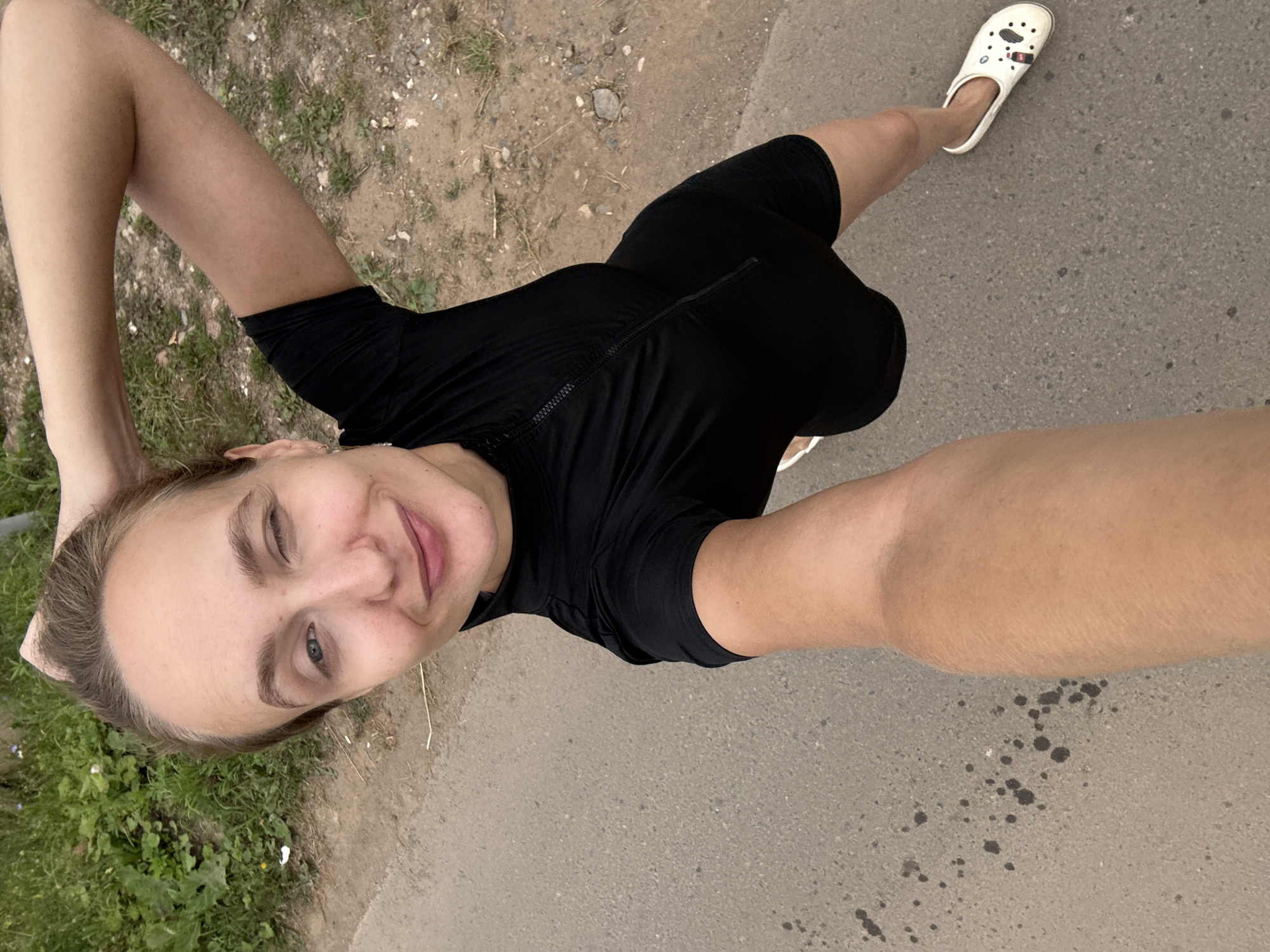 A young woman with blonde hair pulled back is standing on an asphalt road with a grassy area to her left, holding her left arm up behind her head. She is wearing a black short-sleeved athletic outfit with a zipper down the front and white clogs with cartoonish designs. She is winking and smiling at the camera, and her right arm is extended, holding what appears to be a smartphone, suggesting she is taking a selfie. The background shows a dirt and gravel shoulder with sparse green vegetation. The image appears to have been taken outdoors in a rural or semi-urban area.