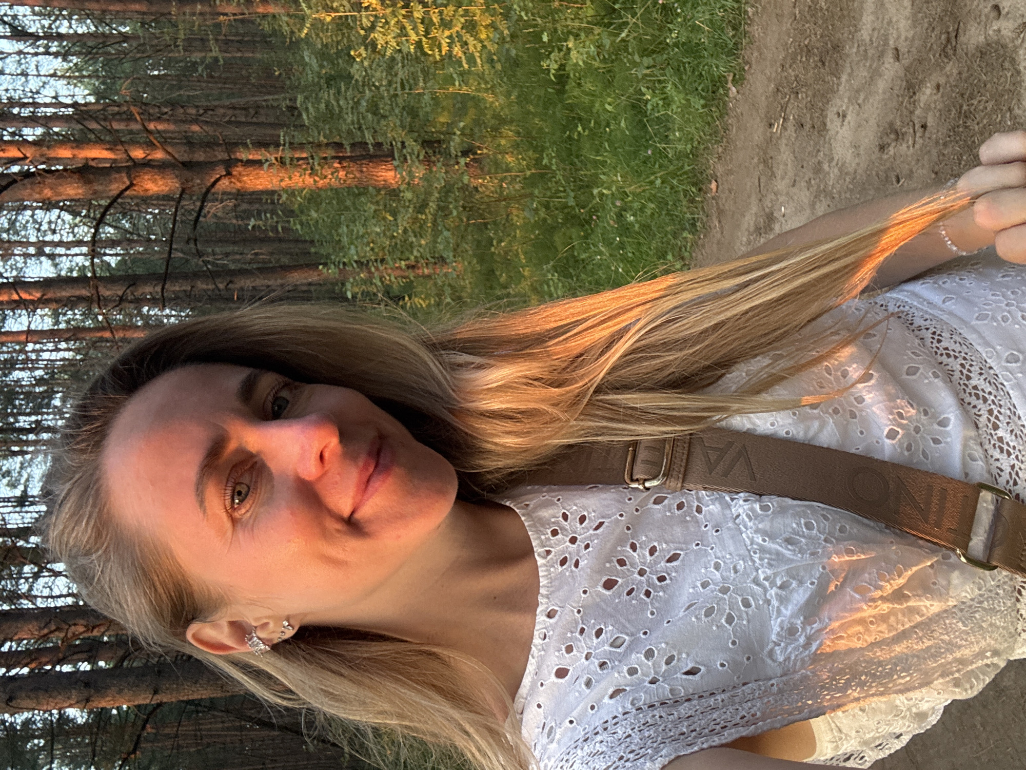 A young Caucasian woman with blonde hair is taking a selfie in a forest. She is wearing a white, eyelet-lace top and a brown crossbody bag. Her left ear is adorned with a cluster of silver earrings. The background is a dense forest with tall trees, and the setting appears to be either sunrise or sunset, as indicated by the warm, golden light illuminating the scene. The woman is standing on a dirt path. The location is not identifiable as Chaikovsky, Russia, or any specific city.