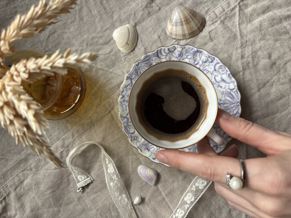 An overhead, close-up shot depicts a tabletop still life arrangement on a light tan, wrinkled linen-like fabric surface. A pale-skinned hand, with a silver-toned ring featuring a white pearl and small clear stones on the ring finger, holds a small white ceramic cup containing dark brown liquid with a thin layer of foam. The cup has a gold rim and is decorated with blue, stylized floral patterns, resting on a matching scalloped saucer. In the upper right quadrant, an amber glass vase contains dried wheat stalks. To the right of the cup, two seashells are visible: one light off-white, and another with brown and tan stripes. On the left side of the frame, a segment of white lace ribbon, with a silver-colored lobster clasp at one end and a single white tear-drop shaped pearl attached to the other end, lies adjacent to a small, smooth, multi-tonal purple and white stone. The scene is stated to be located in Kudrovo, Russia; however, this geographical detail cannot be ascertained from the visual content of the image.