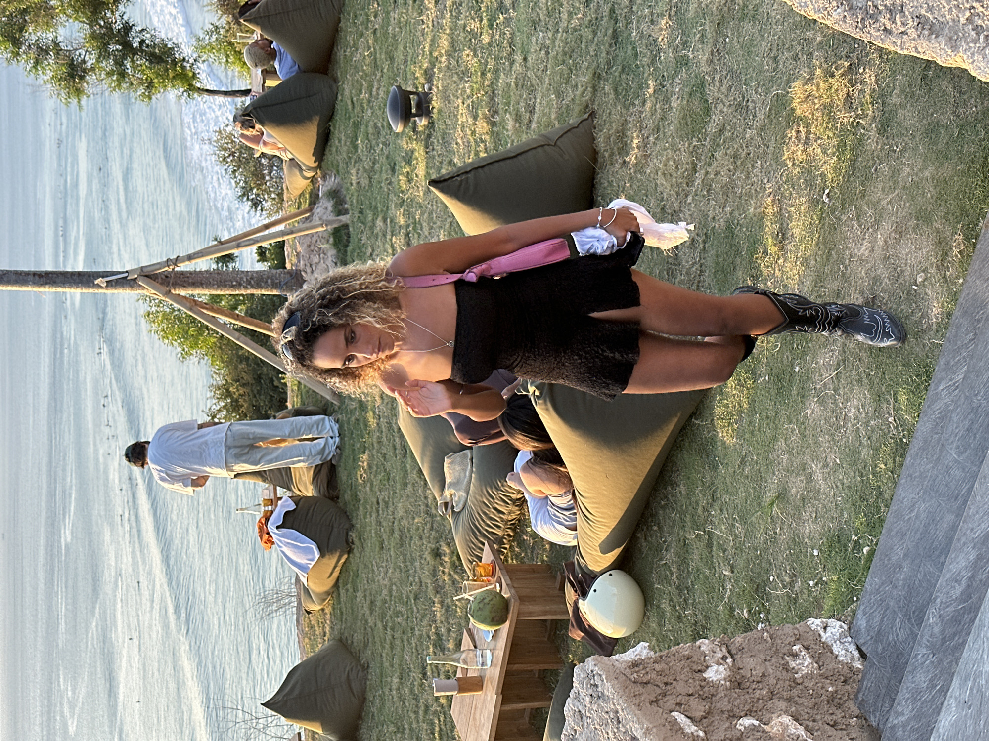 In Kuta Selatan, Indonesia, an outdoor seating area overlooks the ocean where surfers are visible. A woman with curly hair, wearing a black strapless dress and cowboy boots, walks towards the foreground. She carries a pink bag over her shoulder and holds a white cloth in her right hand. To her left, a small wooden table holds drinks and a coconut. In the midground, several large, olive-green beanbag chairs are scattered on a grassy lawn. One beanbag has a person lying on it, facing away from the camera. Further back, a man in a white shirt and light-colored pants stands near a table. The environment suggests a casual, seaside gathering or relaxation spot.