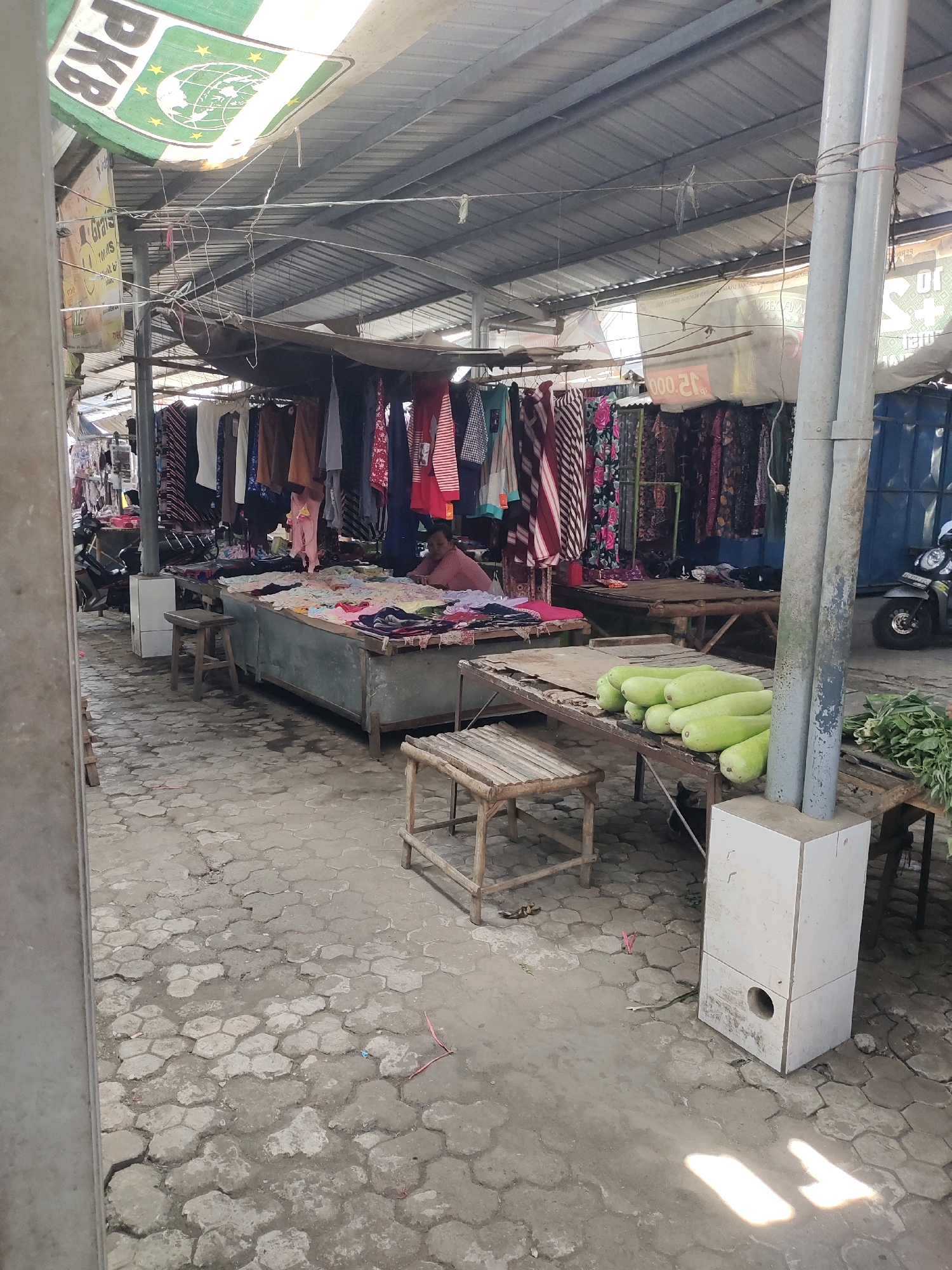 The image depicts a market stall, likely in Sambi, Indonesia, with goods displayed under a corrugated metal roof supported by poles. The ground is paved with interlocking hexagonal concrete blocks. In the foreground, a wooden table holds several green gourds and some leafy green vegetables. To the left, a light-colored, rectangular speaker box rests on the ground. Further back, another stall displays a variety of colorful fabrics and clothing hanging from a rack. A person wearing a pink top is seated behind a table covered with folded garments. The lighting suggests it is daytime, with sunlight casting shadows on the ground. A portion of a motorcycle is visible on the right side of the image, indicating it's an active market.