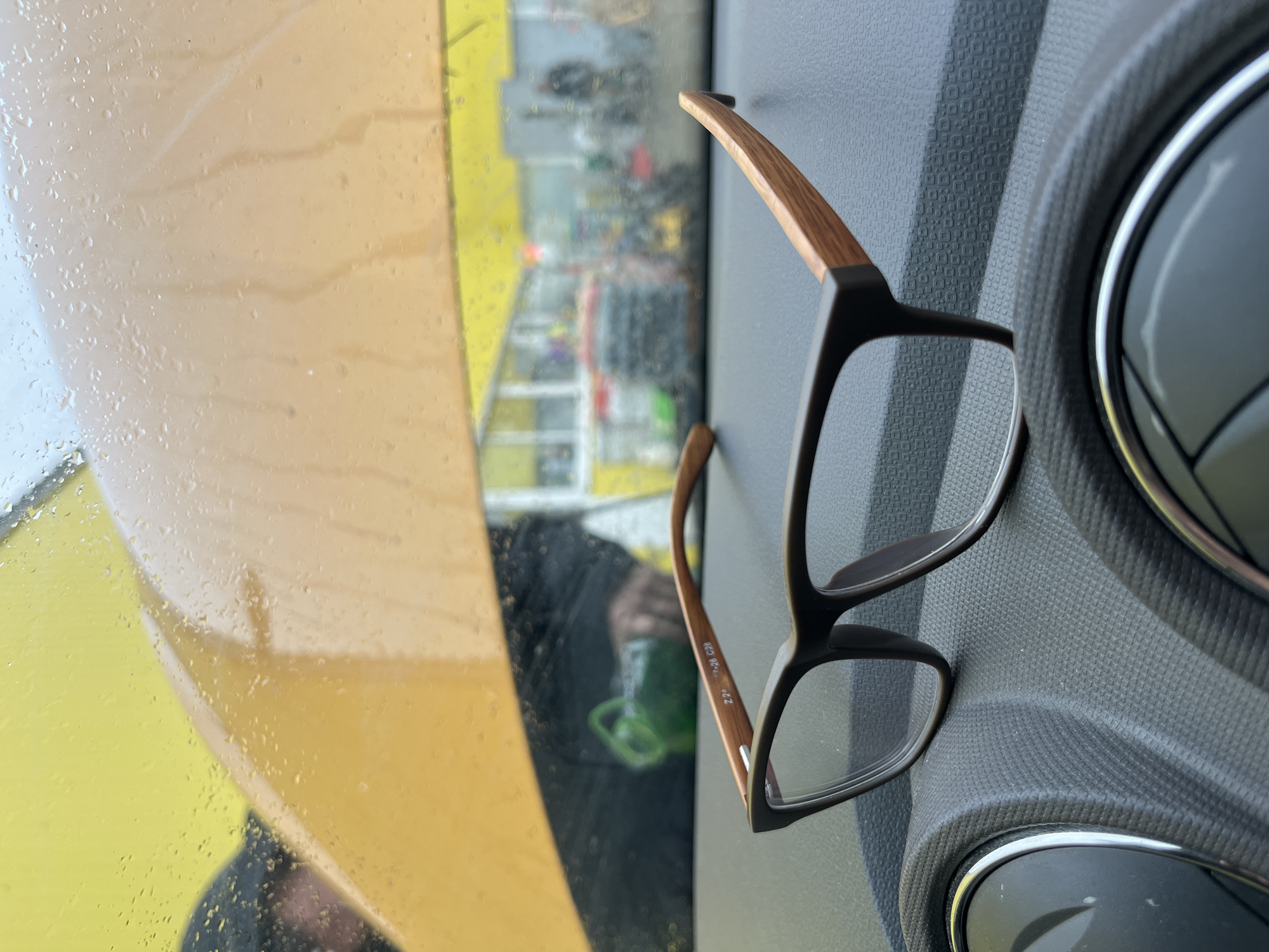 The image shows a pair of eyeglasses resting on a textured gray surface, likely the dashboard of a vehicle. A yellow, rain-streaked window is visible to the left, and through the window, a blurred cityscape can be seen, partially obscured by rain or condensation. The eyeglasses have black frames and wood-grained temples. The reflections in the window suggest the presence of at least one person inside the vehicle, wearing a face mask. The scene appears to be taken during or shortly after rainfall. There is no verifiable evidence in the image to suggest it is in Shevlyakovo, Russia.