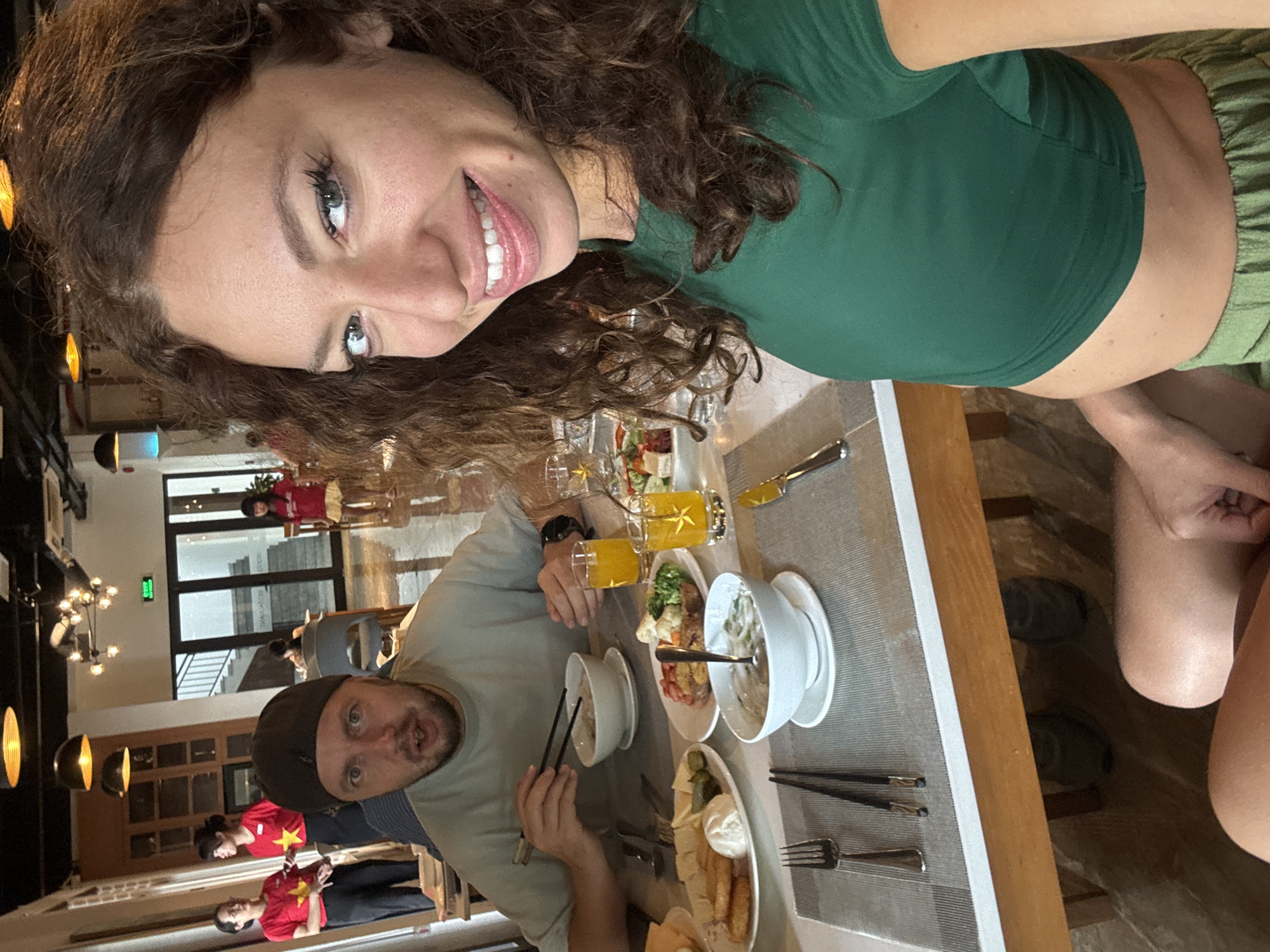 A selfie captures two individuals dining in a well-lit indoor restaurant setting in Đà Nẵng, Vietnam. The woman taking the photo, visible in the foreground with long curly brown hair, wears a dark green crop top and light green shorts, smiling broadly at the camera. Opposite her, a man in a light-colored t-shirt and a dark cap looks towards the camera with a slightly surprised expression, holding chopsticks above a bowl of food.
The dining table is set with various dishes, including bowls of what appears to be soup or noodles, plates with salads and other prepared foods, glasses filled with orange juice, and cutlery. The woman's bare feet are visible under the table.
In the background, the restaurant features modern light fixtures, tables, and chairs. Several other patrons are present, with at least three individuals wearing distinctive red t-shirts featuring a yellow star, characteristic of the Vietnamese flag, serving as a strong location cue. Large windows in the background suggest daylight outside, illuminating the space. The overall atmosphere appears relaxed and social, indicating a meal shared between the two main subjects. A small green sign with indistinct white text is visible on a wall in the deeper background.