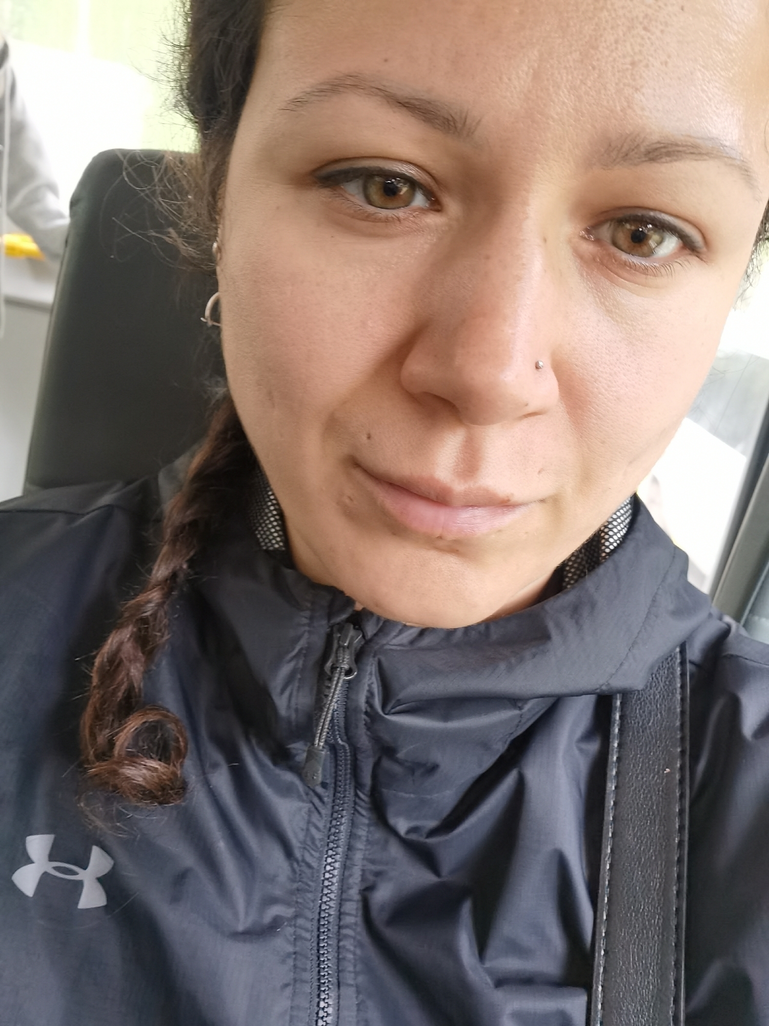 A close-up selfie captures a woman, likely in her 20s or 30s, looking directly at the camera with a neutral, calm expression. Her brown eyes are prominent, and she has dark hair pulled back, with a distinctive braid visible over her left shoulder. A small silver stud pierces her right nostril, and a simple hoop earring adorns her left ear.
She is wearing a dark, possibly black or navy, zippered jacket, which appears to be a windbreaker or light sports jacket. The jacket features a visible light gray "Under Armour" logo on the left chest area. Underneath the main jacket collar, a section of patterned fabric, possibly a checkered or dotted lining, is visible. A black strap, likely from a bag, crosses her right shoulder.
The background is softly blurred, suggesting a shallow depth of field. To her left, a blurry figure in a light-colored hooded garment is partially visible, hinting at another person nearby. To her right, a bright, indistinct area suggests a window or open space, indicating an indoor or vehicle setting during daytime. No specific cues directly reference Chaikovsky, Russia.