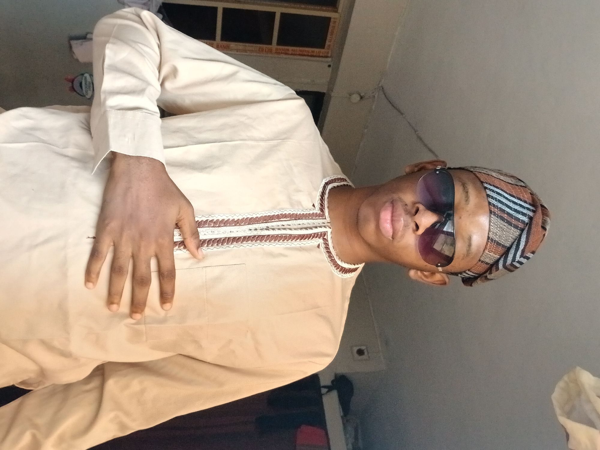 A young Black male, appearing to be in his late teens or early twenties, is captured in a medium-shot selfie taken indoors. He is wearing a traditional light beige buba, a long-sleeved tunic, adorned with a decorative brown and white patterned trim around the neckline and down the center front. He also wears a matching patterned cap, possibly a fila, on his head. His face is partially obscured by dark aviator sunglasses. His right hand is placed flat across his chest, just below the sternum. The background suggests a modest urban dwelling; a window with vertical blinds is visible to the left, and to the right, clothing is hung, possibly on a clothesline or rack. The lighting suggests an indoor environment, likely during daylight hours, casting soft shadows. The overall impression is of a posed portrait, possibly in a residential area within Lagos, Nigeria, given the cultural attire.