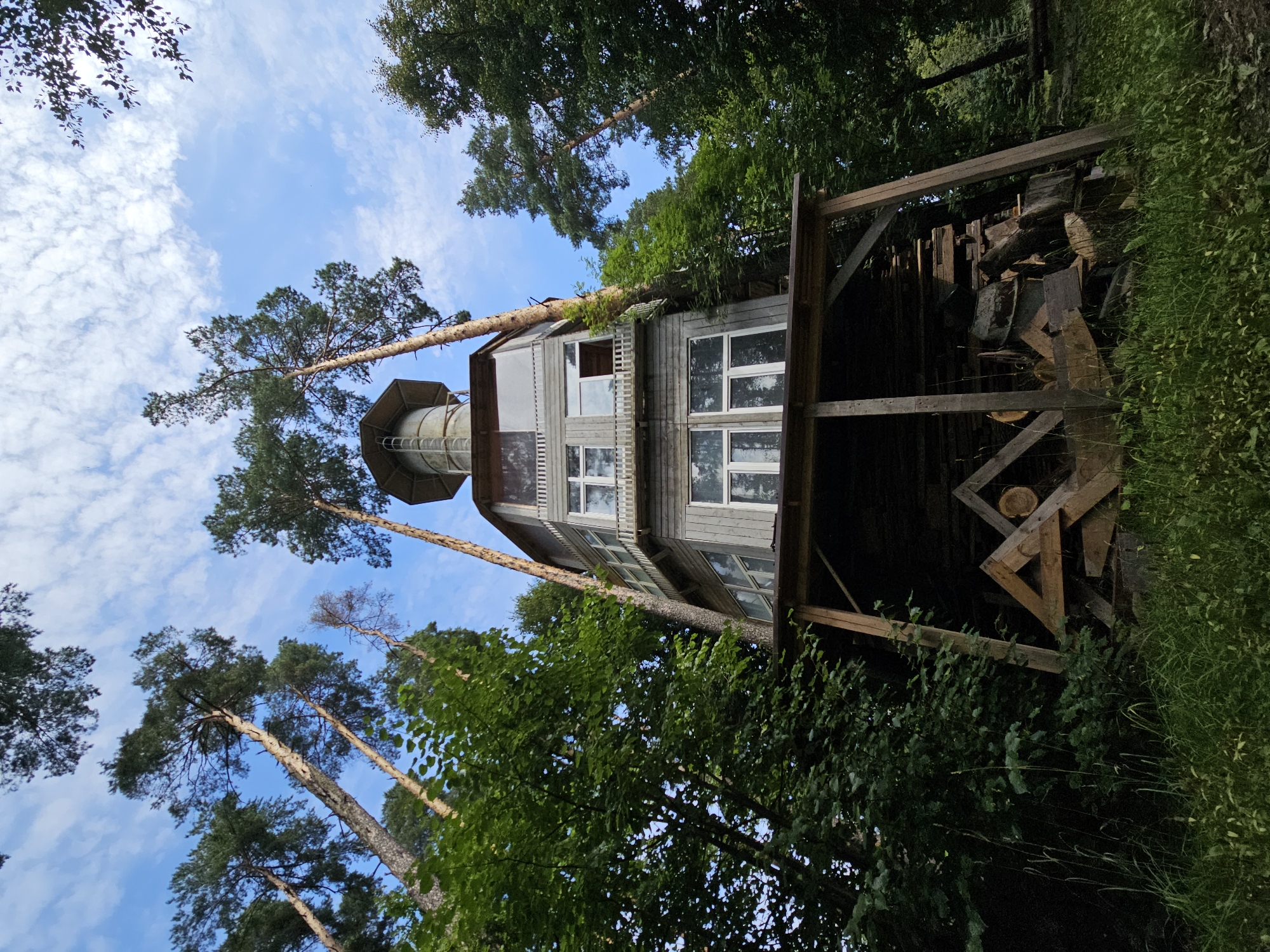This is a low-angle, full shot of a unique, multi-story wooden structure resembling a tower or lighthouse, set amongst tall pine trees. The building has a cylindrical base and octagonal upper sections with numerous windows and balconies. A water tower-like structure tops the building. The structure is built on stilts, supported by wooden beams. Below the main structure, there is a sheltered area with stacked lumber and other wooden materials. The surrounding environment is a dense forest with lush green foliage and tall trees reaching towards a bright blue sky with scattered white clouds. The image was taken in the village of Ulitkino, Russia. There are no people visible in the image.