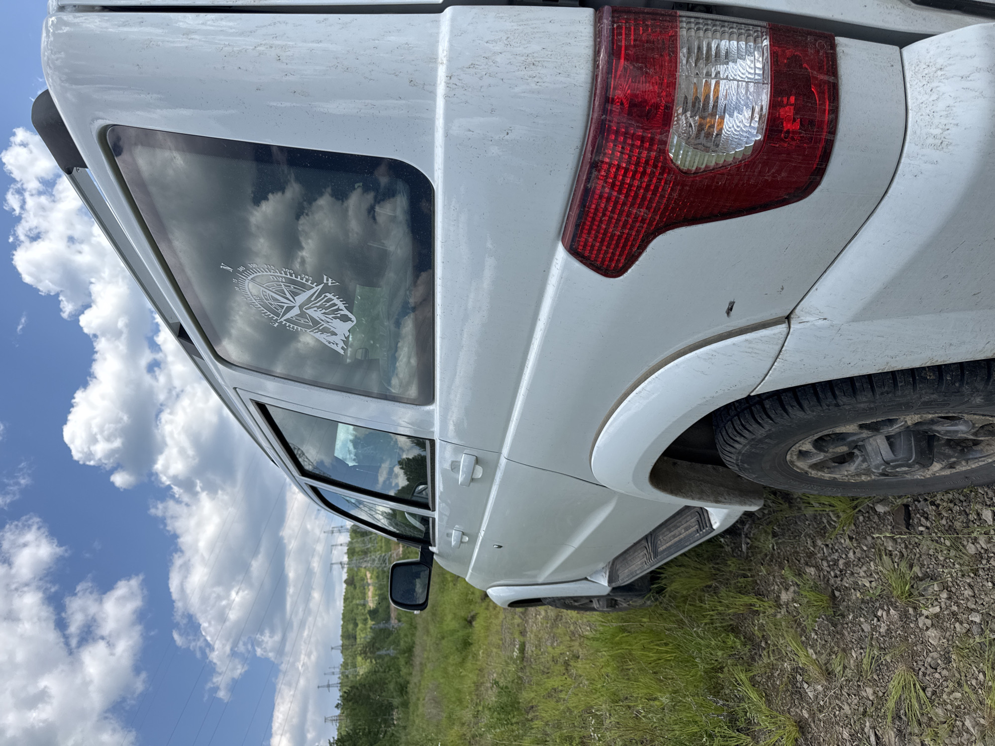 This is a close-up, side-view, low-angle shot of the rear quarter panel of a white, dirt-splattered SUV parked on a gravel and grass embankment. The vehicle is facing to the left of the frame, with its back end towards the right. The sky, filled with fluffy white clouds against a bright blue backdrop, is reflected in the tinted rear window, which features a white sticker of a compass rose with mountains and a wolf's head. The SUV's red taillight assembly is prominent on the right side of the image, showing a clear lens on top and a red housing below.
The setting appears to be an outdoor, rural location, possibly in Skovorodino, Russia, given the context. The ground is covered with a mixture of dirt, gravel, and green grass. In the background, beyond the vehicle, a line of green trees stretches horizontally, and further in the distance, power lines and transmission towers are visible against the sky, suggesting proximity to an area with electrical infrastructure. The lighting and cloud patterns indicate it is daytime, likely with fair weather. The vehicle's dusty condition suggests it has been used in off-road or unpaved environments. No people are visible in the image. There is no discernible text on the vehicle or in the environment, other than the details on the compass sticker.