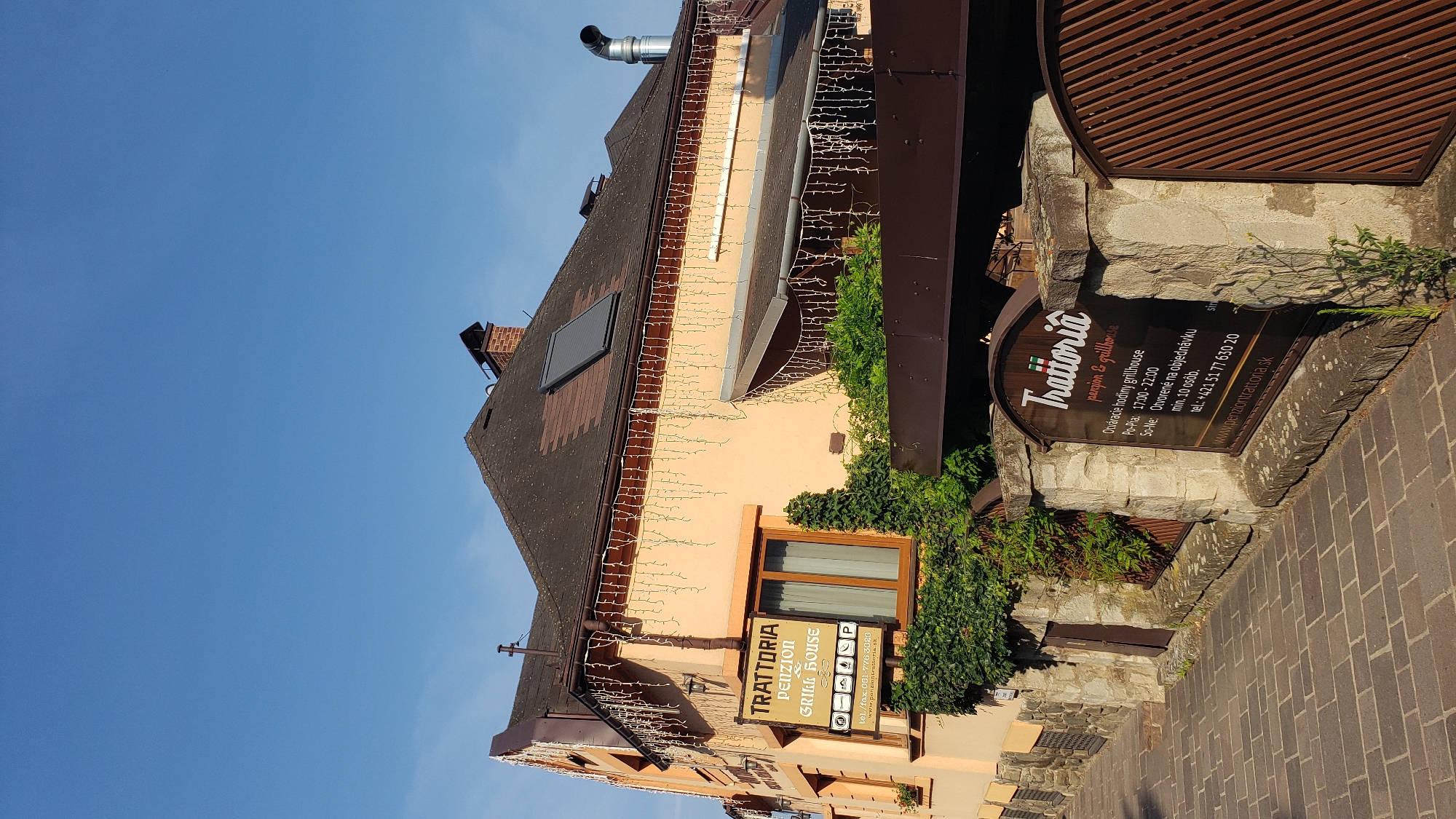 The image shows a two-story building with an orange facade, a brown tiled roof, and decorative icicle lights hanging from the eaves. The building has a solar panel on the roof and two chimneys. A sign on the front of the building reads "TRATTORIA PENZION & GRILL HOUSE". To the right of the building, a stone wall with a dark wooden sign is visible. This sign also reads "Trattoria penzion & grillhouse" and lists operating hours and a phone number. A cobblestone path leads up to the building. The sky is clear and blue. The scene depicts the entrance to a restaurant and guesthouse in Prešov, Slovakia.
