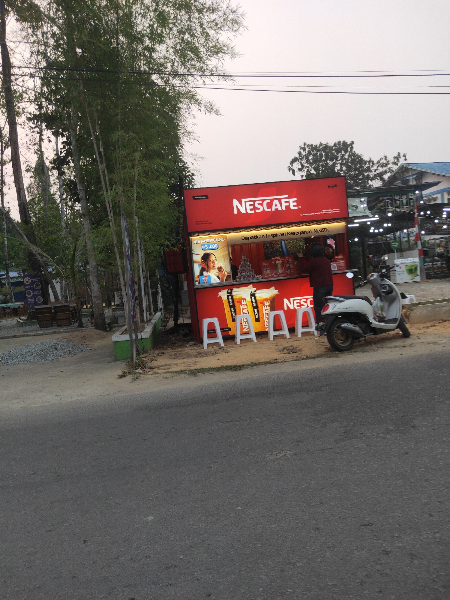 A Nescafé branded kiosk is set up on the side of a road in Sintang, Indonesia. The kiosk is red and features prominent Nescafé logos and promotional material. A large digital display shows a woman with headphones holding a cup. Several white plastic chairs are arranged in front of the kiosk, and a white scooter is parked to the right. The scene is flanked by tall trees and vegetation, with a street in the foreground. The sky is overcast, suggesting it might be early morning or late afternoon. The overall impression is of a street-side business operating in a tropical environment.