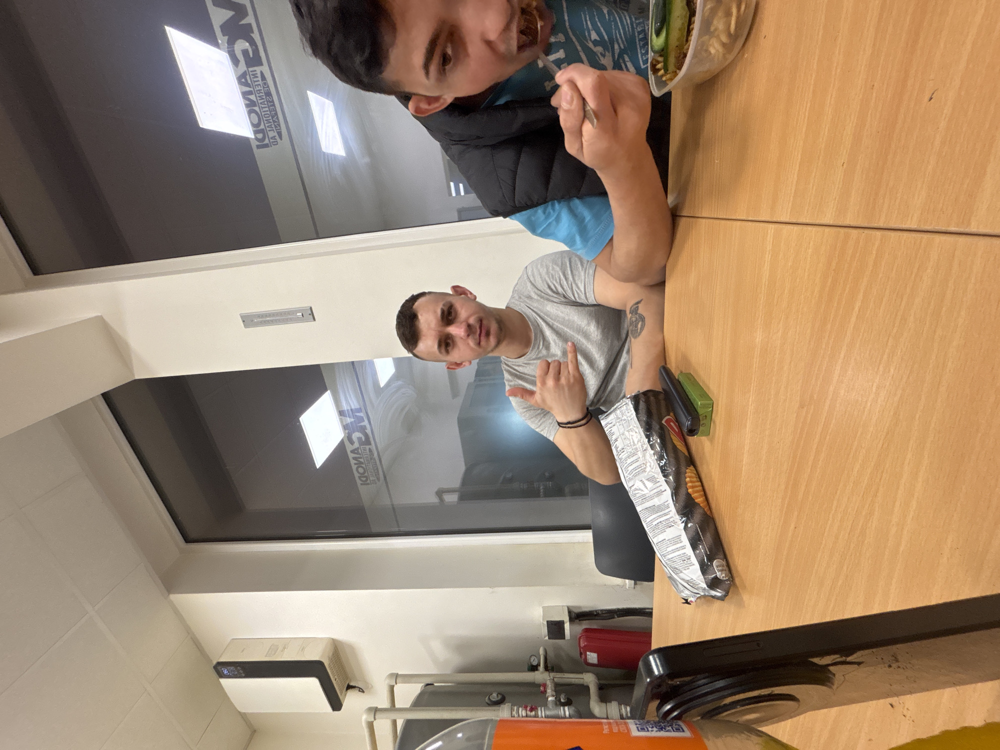 A photograph depicts two men seated at a light brown wooden table indoors. One man is eating from a clear plastic container using a utensil. He is wearing a blue shirt and a black vest. The other man is looking at the camera and making a hand gesture. He is wearing a grey shirt. A partially consumed bag of chips rests on the table next to the man in the grey shirt. In the background, a window with a corporate logo is visible, and below it, there are pipes and equipment. The location is in the city of Unknown, Unknown.