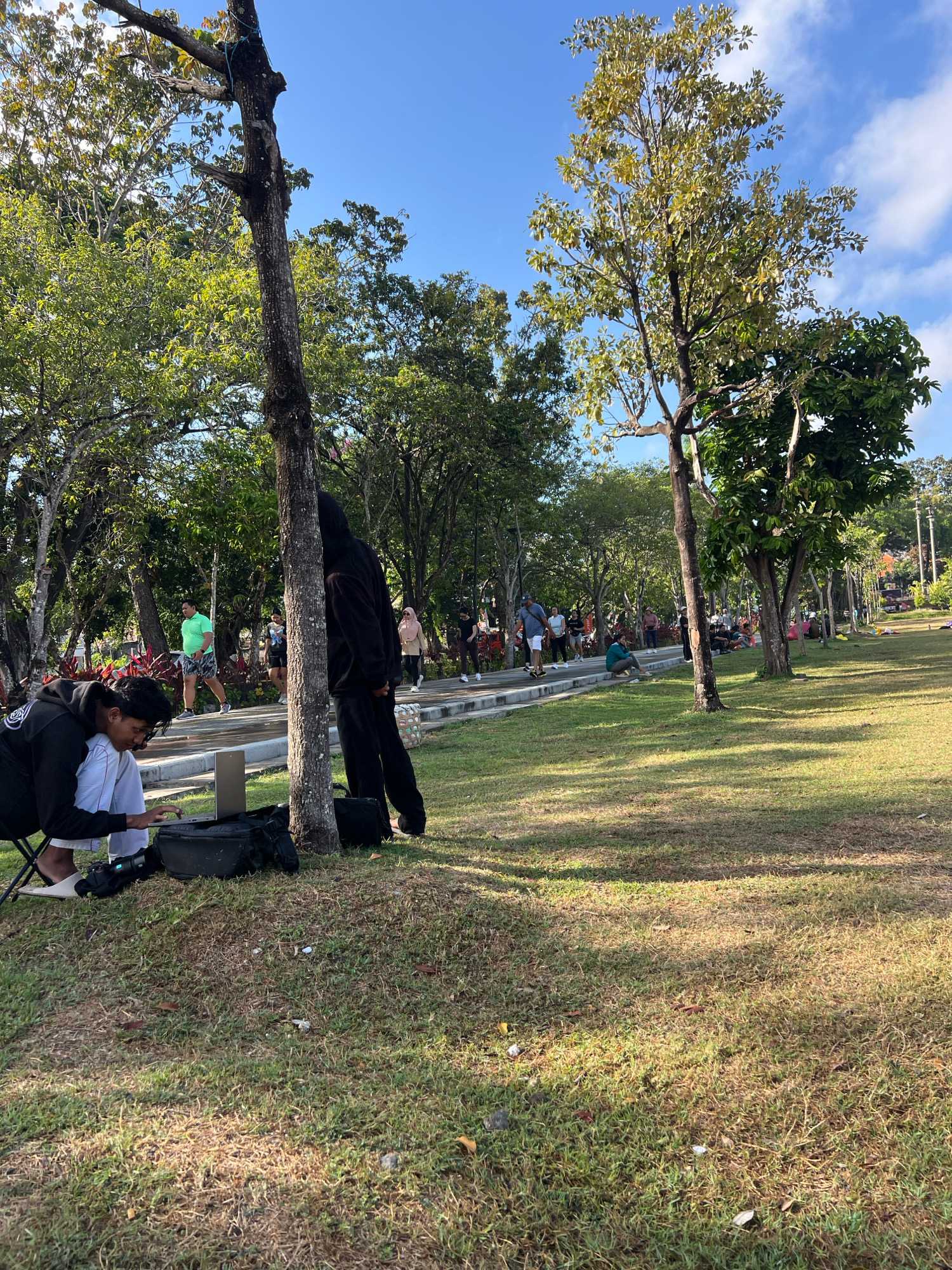 The image depicts a sunny day in a park, possibly in Denpasar, Indonesia, with a clear blue sky and scattered clouds. In the foreground, two young men are present. One is seated on a folding chair, wearing a black hoodie and white pants, and appears to be interacting with a laptop placed on a black bag. Next to him, another person, also in a black hoodie and black pants, stands leaning against a tree. Their face is obscured by the hoodie. A camera is also visible on the grass next to the seated individual.
The background of the park shows a paved walkway with several people engaged in various activities such as walking and conversing. Trees with lush green foliage are scattered throughout the park, casting shadows on the well-maintained grassy areas. The overall atmosphere suggests a recreational setting where people are enjoying outdoor activities.