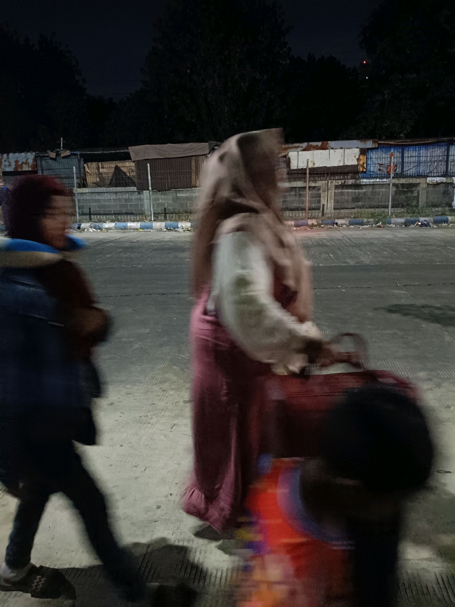 This is a nighttime, slightly blurred photograph taken in Kedungrejo, Indonesia. In the foreground, three figures are moving. On the left, a person wearing a blue jacket over a red shirt is visible from the chest down, with their legs and feet in motion. They are wearing sandals with a zigzag pattern. To the right, a person in a light brown hijab and a long, dark pink garment is walking, carrying a dark red bag with a strap in their right hand. Partially obscuring the central figure is a third person, who appears to be a child, moving from right to left. This child is wearing a red and blue garment and has dark hair. The background consists of a concrete surface that appears to be a road or parking lot, with a low concrete barrier along the edge. Beyond the barrier, there are several small, makeshift structures with corrugated metal and wooden elements. Dark trees line the far background, and the sky is dark. The lighting suggests artificial illumination from the side, casting shadows.