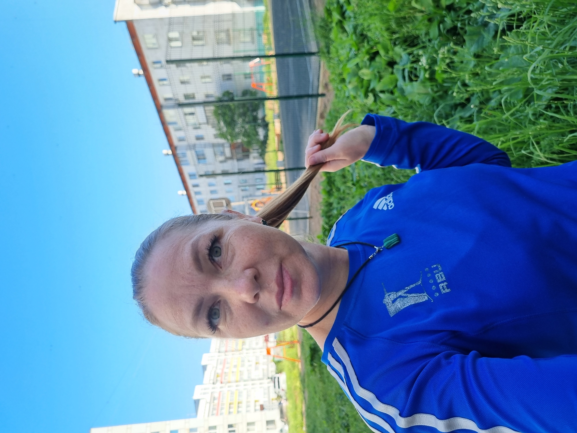 In a full-frame, eye-level, outdoor selfie, a Caucasian woman with blonde hair pulled back, is captured. She is wearing a blue athletic shirt with white stripes on the sleeves and a necklace with a green pendant. The woman is holding a strand of her hair. The background shows a residential area in Chaikovsky, Russia, with apartment buildings and a fenced-in sports court. The sky is clear and blue. The foreground is filled with green grass.