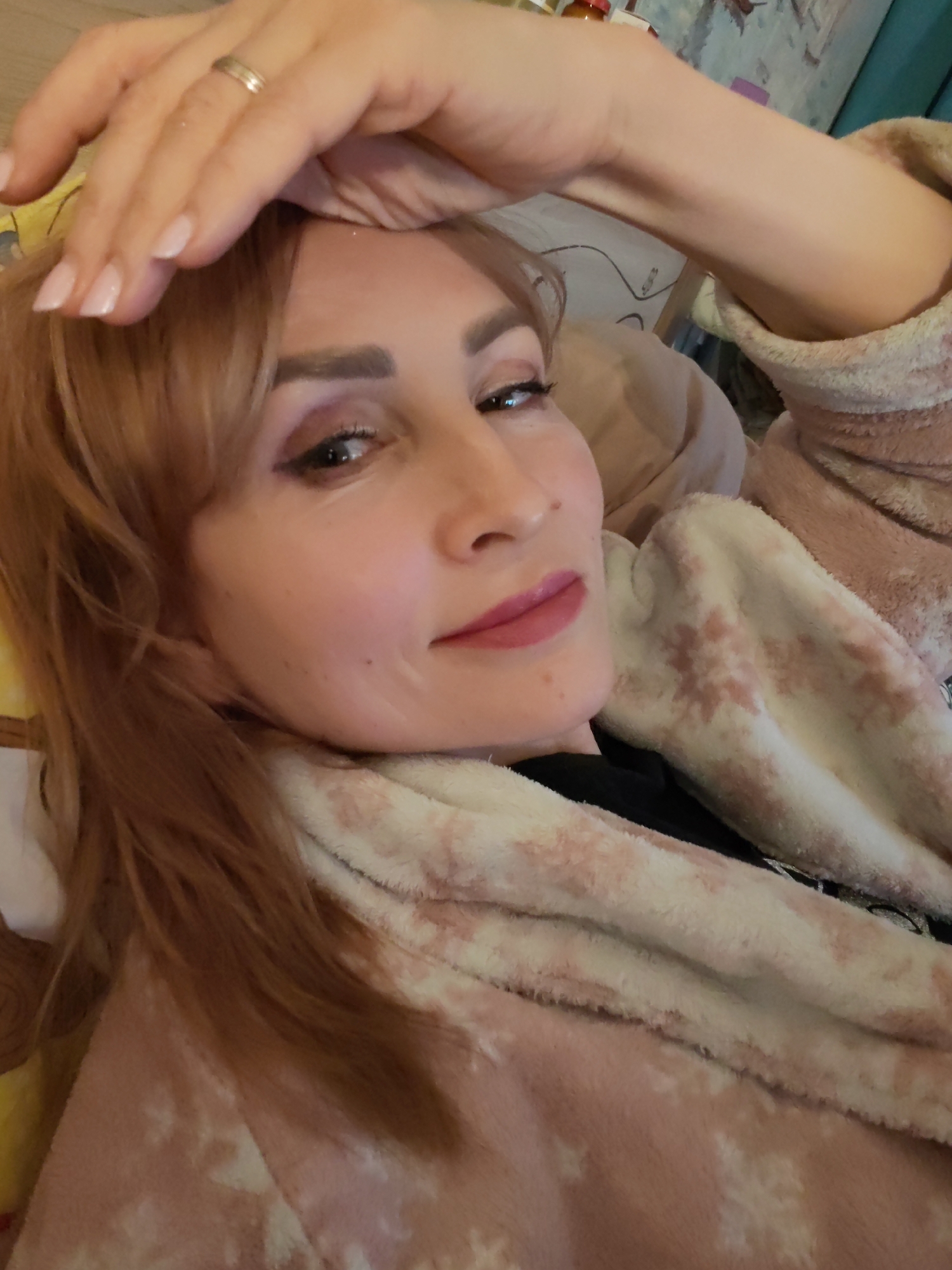 A woman with light brown to auburn hair is captured in a close-up selfie, looking directly at the camera with a slight, relaxed smile. She is lying down, with her left hand resting on her forehead, revealing manicured nails with a light polish and a simple ring on her ring finger. Her makeup includes winged eyeliner, shimmering eyeshadow, and pink lipstick. She appears to be wearing a fluffy, light pink and white patterned robe or blanket, which covers her body and arms, suggesting a cozy and comfortable setting. A black garment is visible underneath the robe near her chest.
The background is softly blurred but indicates an indoor environment, possibly a bedroom or living room, with hints of wallpaper or a textured wall and some indistinct objects on what might be a shelf or nightstand. The lighting is soft and even, suggesting an indoor setting without strong natural light. The overall mood conveyed is one of relaxation and calm. There is no visible text in the image. The location context for this scene is Chaikovsky, Russia.