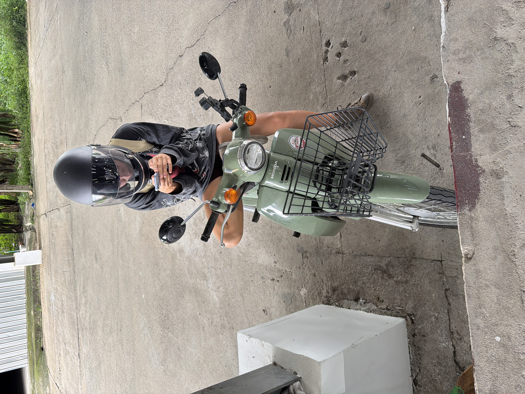 A person sits on a light green Honda Cub motorcycle. The person wears a black helmet with a reflective visor and a dark grey hoodie. They hold a smartphone in their right hand. The motorcycle has a black basket attached to the front. The scene takes place on a concrete surface, likely a street or driveway, with some greenery visible on the left side of the image. The sun is out, casting shadows, suggesting it is daytime. The setting appears to be an outdoor area in Ban Buak Khrok North, Thailand. The image is taken from a high-angle perspective, looking down at the person and the motorcycle.