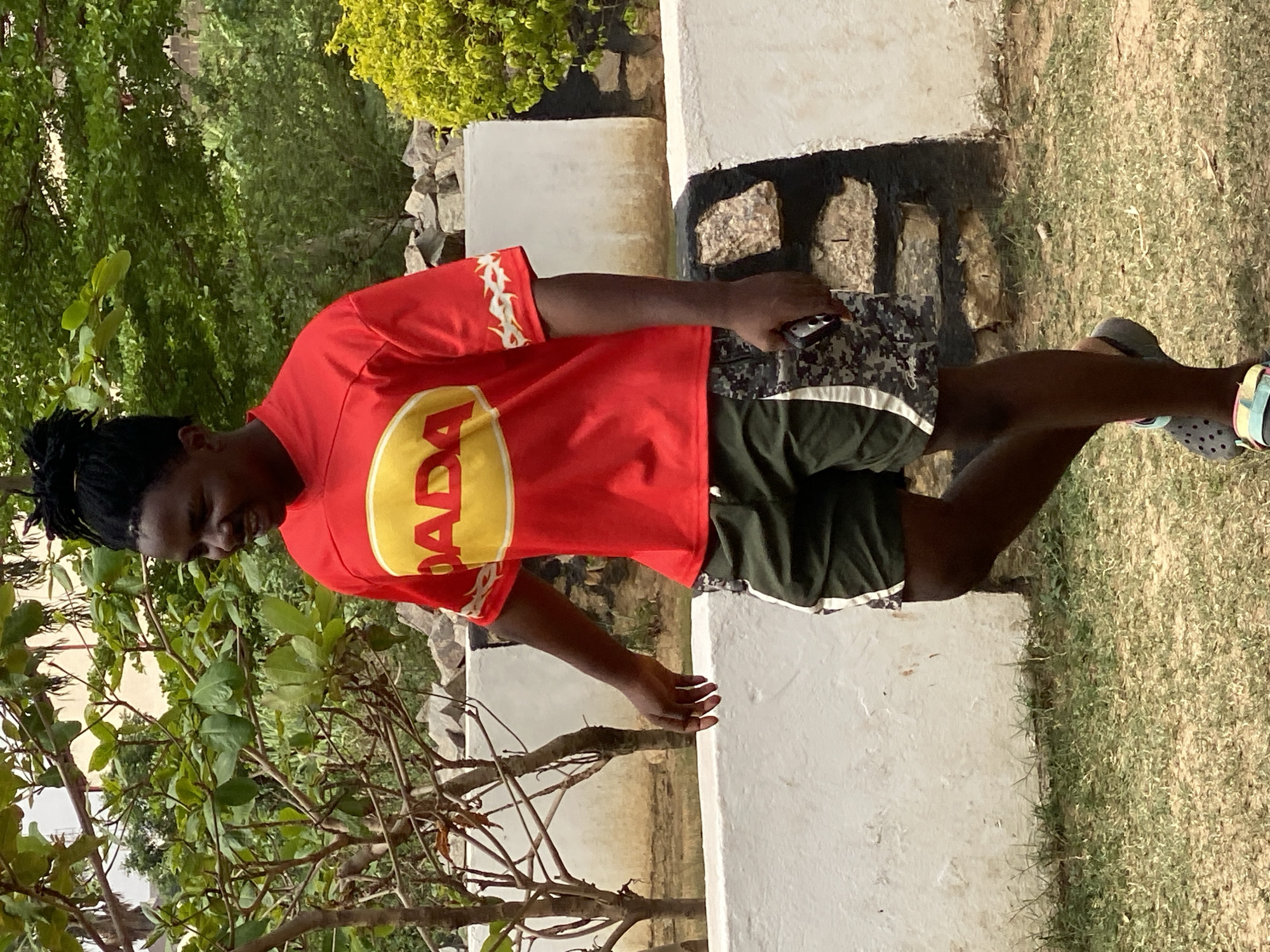 A person stands outdoors, possibly in a residential or public green space in Tundun Wada South, Nigeria. They are wearing a red t-shirt with a yellow circular logo and dark-colored shorts. The person's footwear includes open-toed shoes. The background features trees, grass, and what appear to be low concrete walls or planters. The person is holding a small object in their right hand.