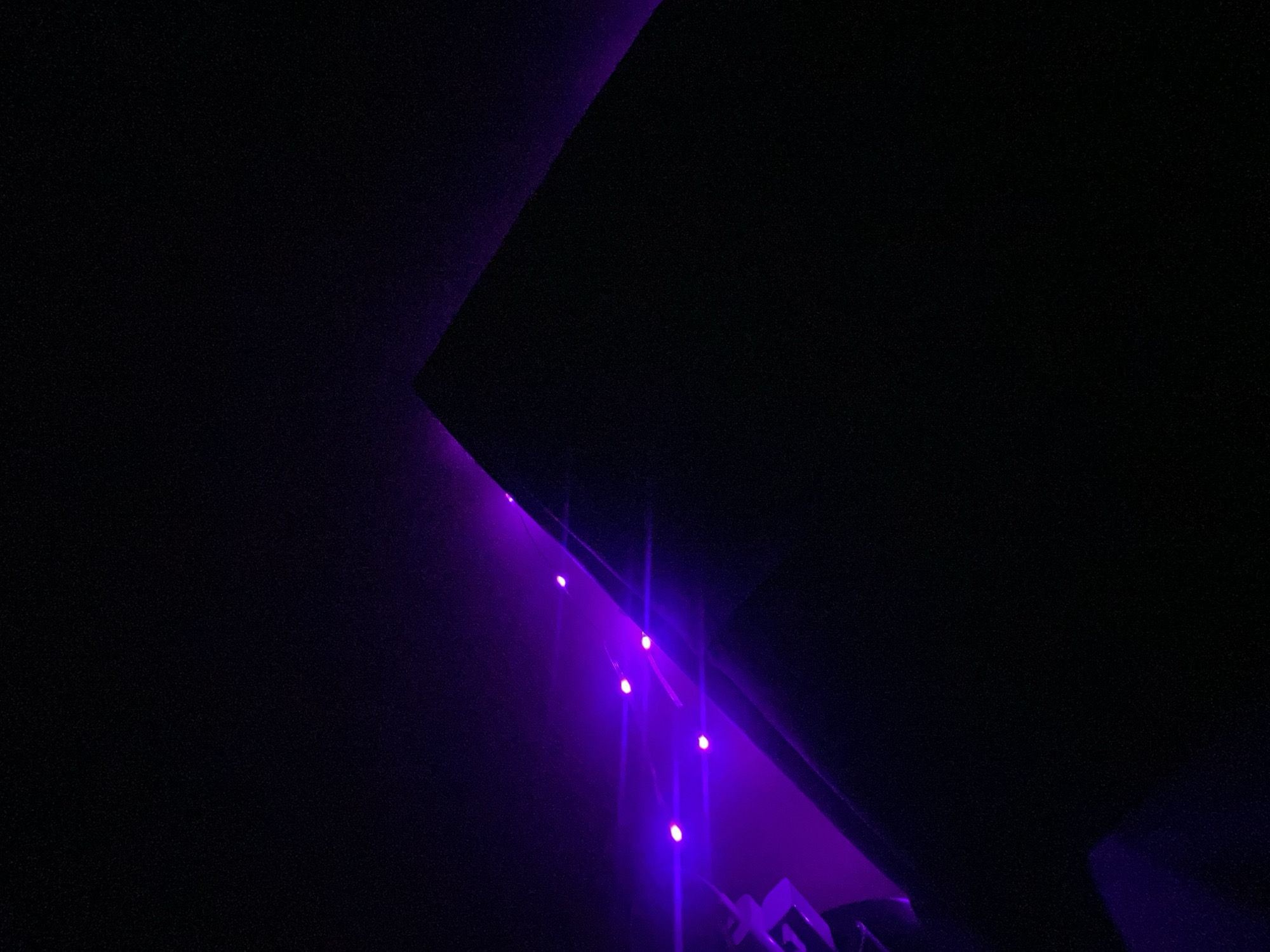 The image shows a dark, indoor setting in Yekaterinburg, Russia. The upper portion of the frame is dominated by dark, angular surfaces. A row of purple lights is visible along the edge of one of the surfaces, emitting streaks of light downwards. Partial views of metallic objects are visible at the bottom.