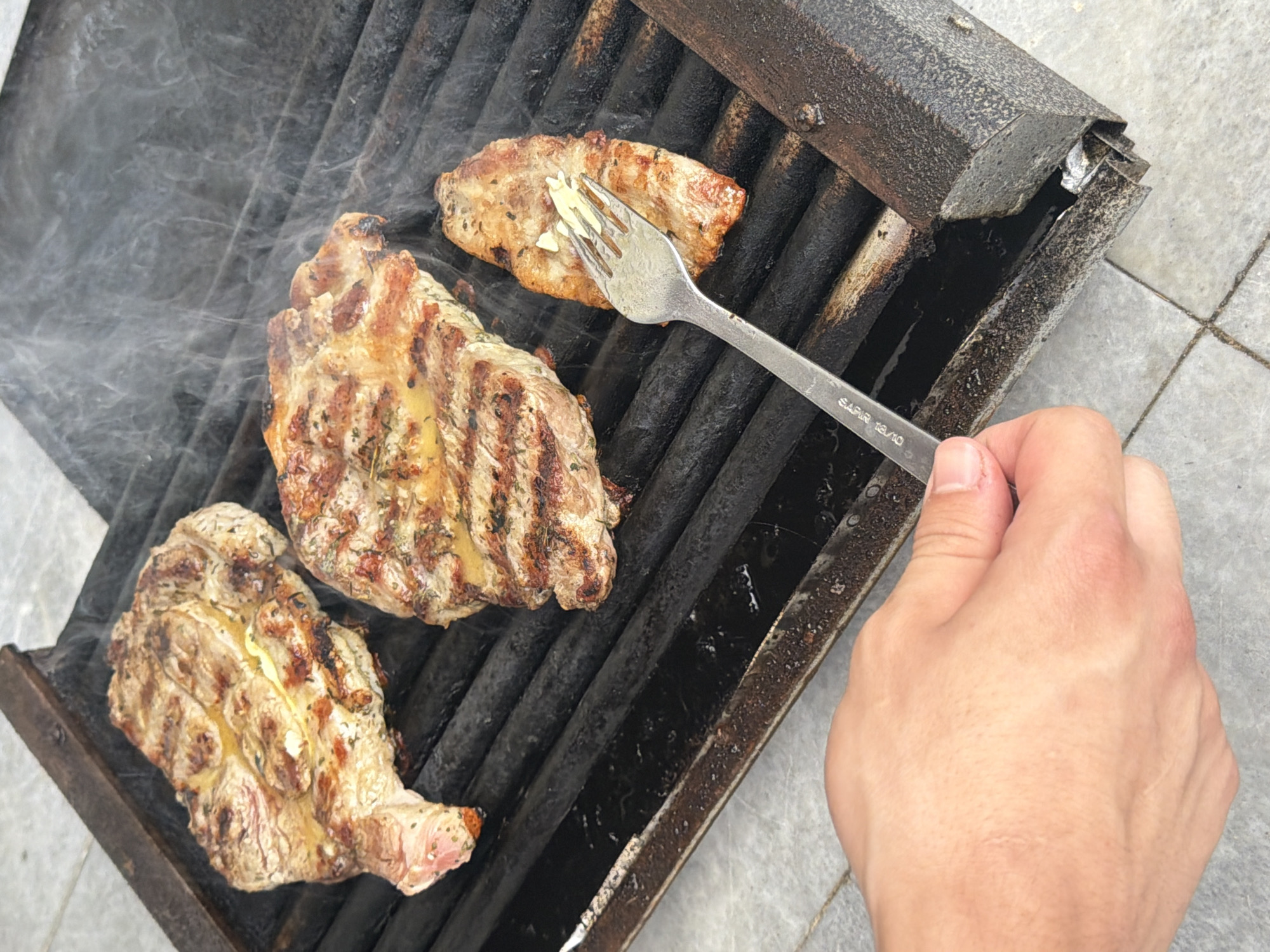 In the city of Svilengrad, Bulgaria, on a barbecue grill, three pieces of seasoned steak are being cooked. The grill is made of black metal and is placed on a light-colored tiled surface. Smoke rises from the steaks, indicating they are actively cooking. A fork, held by a person whose hand is visible in the foreground, is in contact with the smallest steak, suggesting it is being tested for doneness or repositioned. The steaks have grill marks, and some appear to have butter or seasoning on them. The overall scene depicts a moment of outdoor cooking.