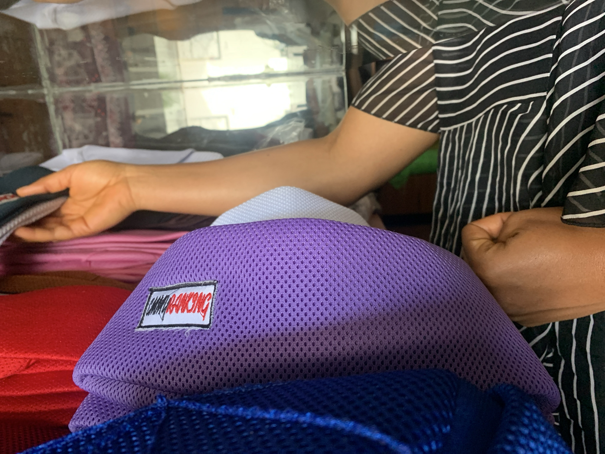 In this medium shot, a person wearing a black and white striped garment is reaching into a display of colorful fabrics. The person's hand is visible, lightly touching a stack of pink fabric. In the foreground, a prominent purple mesh object is seen, possibly a bag or cushion, with a small white label featuring red lettering that reads "CHAIRONG". To the left of the purple object, a red fabric is visible, and below it, a blue mesh object. The background appears to be a reflective surface, possibly glass, showing a blurred reflection of shelves filled with more items. The lighting suggests an indoor setting, possibly a shop or market stall, with bright, diffused light. The scene conveys a sense of browsing or selecting items, with no strong emotional cues evident. The presence of various fabrics and the way they are displayed suggest a retail environment. The visible text on the label is "CHAIRONG".