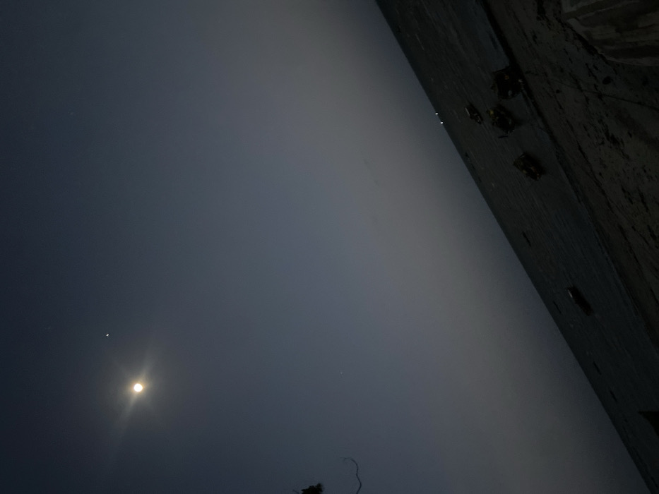 The image displays a tranquil nighttime scene, likely along a coastal or riverside area in Bagamoyo, Tanzania. The dominant feature is a bright, luminous moon positioned in the lower-left portion of the dark sky. To the upper-left of the moon, a distinct, bright star or planet is visible. The sky transitions from a deep indigo-black at the top and left to a lighter, hazy grey across the horizontal middle of the frame, suggesting atmospheric mist or distant light pollution near the horizon.
On the far right side of the image, a dark, textured wall or rocky bank extends vertically, appearing silhouetted against the hazy backdrop. Several indistinct, darker shapes are discernible on or near this structure, possibly representing small boats, debris, or other objects. A faint, skeletal tree branch is barely visible in the very bottom-left corner, silhouetted against the moonlit sky.
The scene depicts nighttime conditions with a clear upper sky contrasting with a misty or foggy layer near the horizon. No explicit activity is taking place, and there is no visible text.