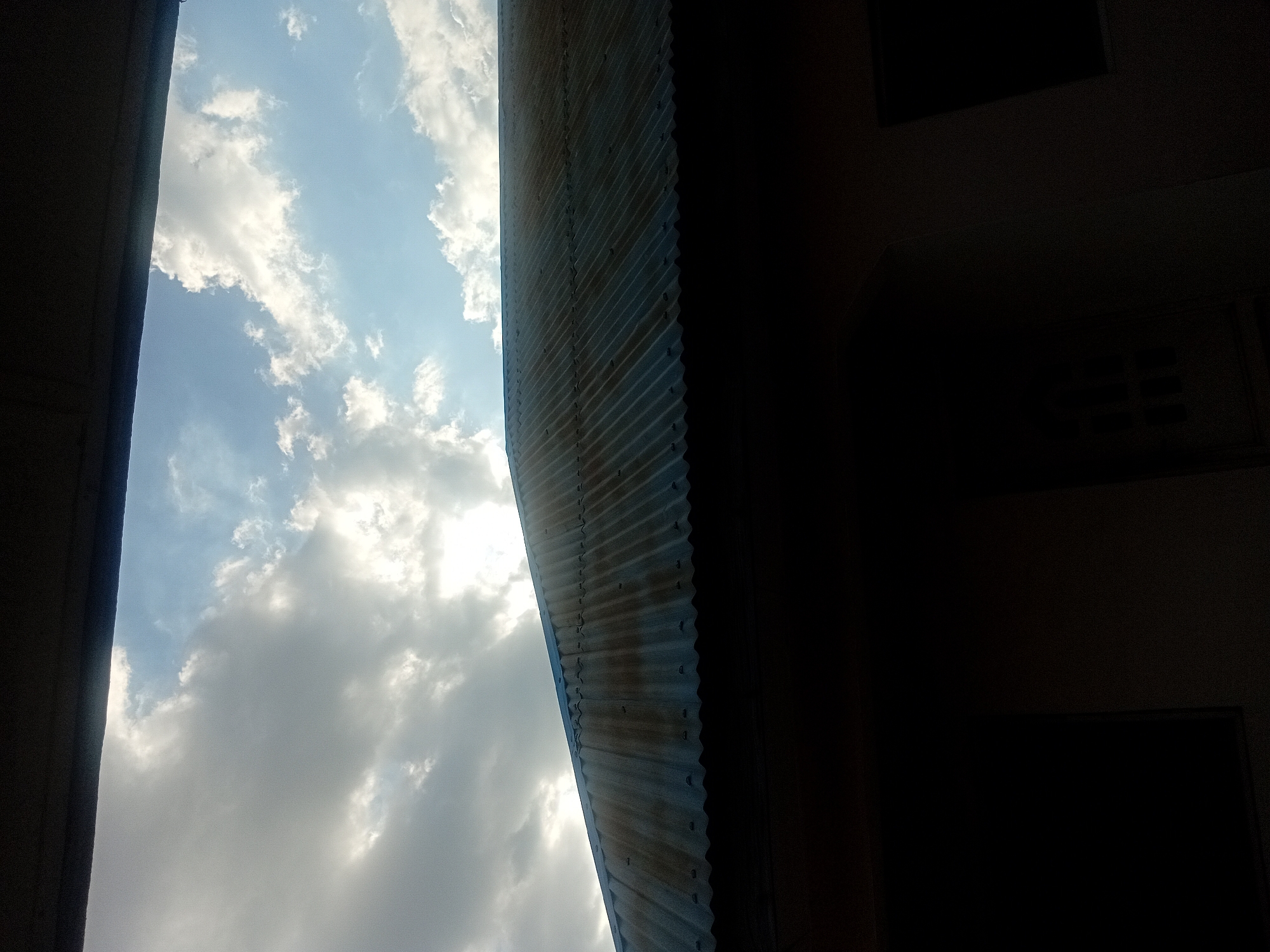 The image, oriented vertically, captures a scene looking upwards towards the sky and the edge of a building. The left half of the frame is dominated by a bright blue sky filled with a mix of white, puffy clouds and darker, greyer clouds, indicating a partly cloudy day. The bright illumination suggests daytime, likely mid-morning or afternoon, with strong sunlight.
A corrugated metal structure, possibly a roof or awning, runs diagonally across the central part of the image, separating the bright sky from a heavily shaded area. This structure features distinct parallel ridges and visible fasteners (screws or bolts), with some signs of discoloration or rust on its surface.
The entire right side of the image is extremely dark, almost black, representing a deep shadow or an underexposed interior space. Within this dark expanse, faint outlines become discernible with close inspection. In the upper right quadrant, a subtle rectangular shape might indicate a window or an opening in a wall. Further down, near the bottom right, some very faint horizontal lines suggest another architectural feature, possibly a vent or louvers. The overall impression is of viewing the sky and the exterior of a structure from within a shaded enclosure.
No specific activity or event is taking place, and there are no people visible. No text is present in the image. The photo was taken in Lokoja, Nigeria.