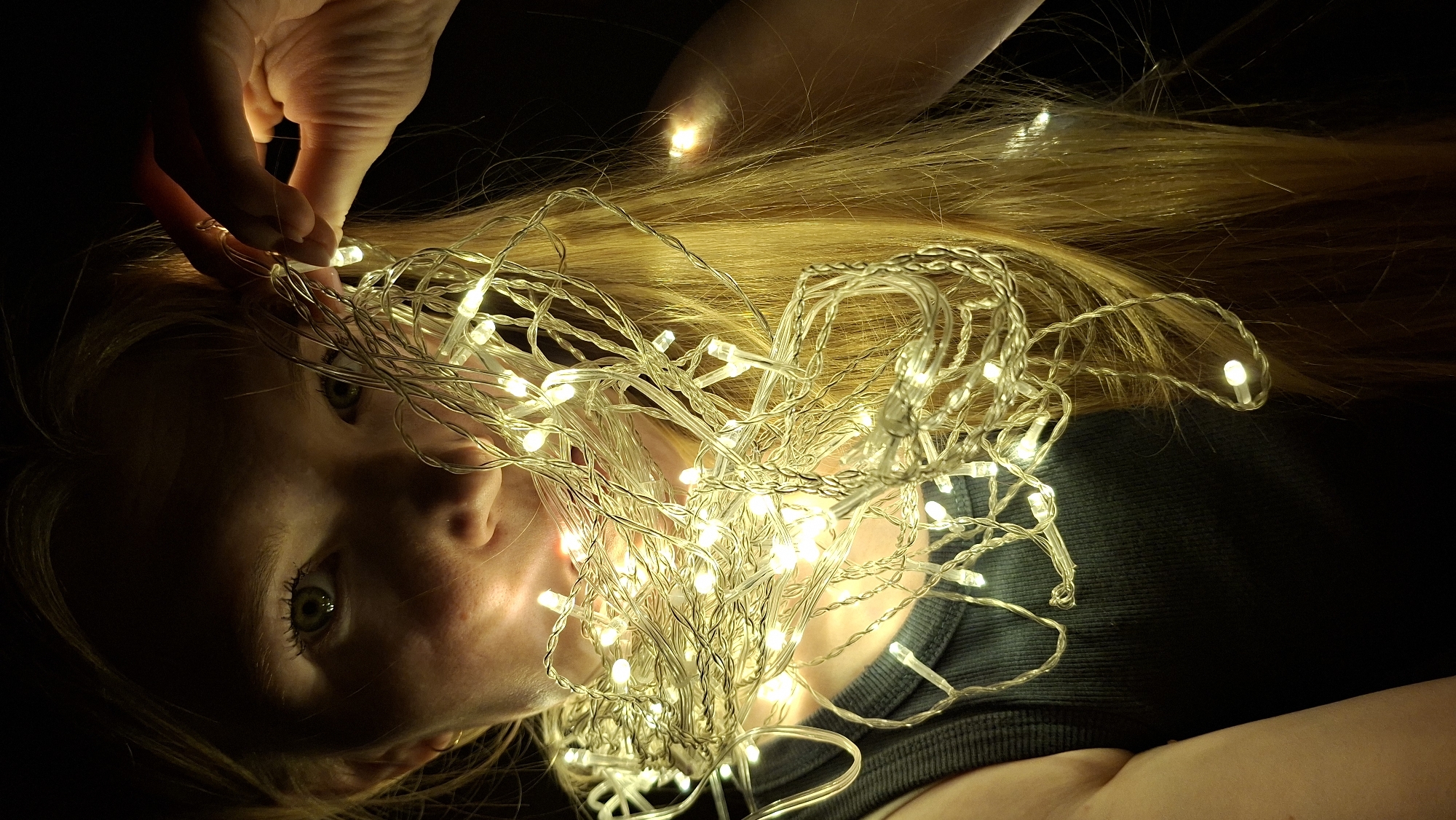 A person is lying on their back in the dark, with their head surrounded by strands of small, glowing fairy lights. The person has fair skin and light blonde hair. One hand is visible near their forehead, seemingly arranging or holding the lights. The background is black and indistinct. There is no evidence to suggest the image was taken in Большой Букор, Russia.