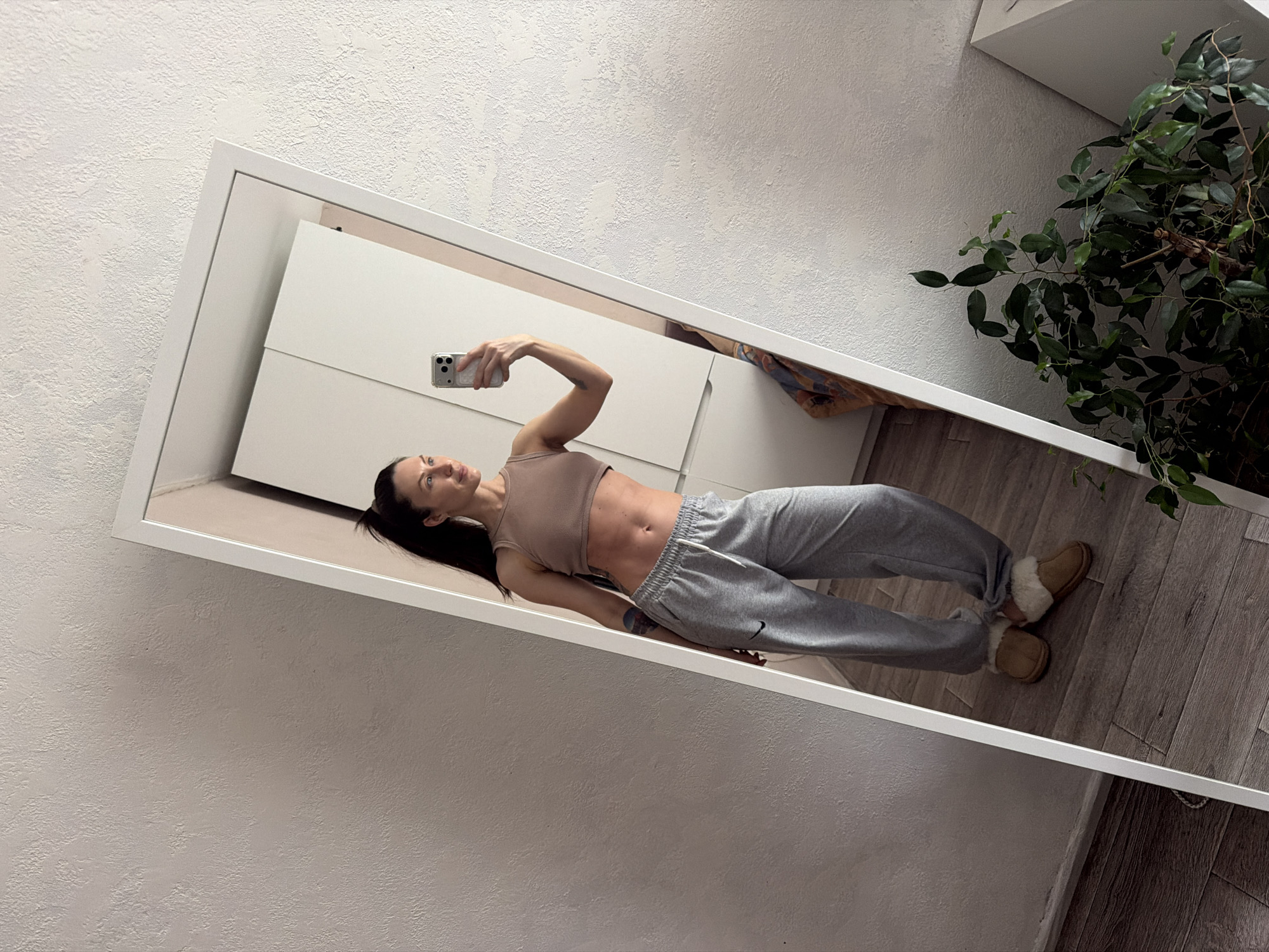 A woman stands in front of a full-length mirror, taking a selfie with her cell phone. She is wearing a light brown crop top and grey Nike sweatpants, along with tan-colored slippers. The setting appears to be inside a residence, featuring a white wall with textured surface, a white cabinet, and wooden flooring. A plant is visible in the corner of the room. The image was likely taken in Perm, Russia.