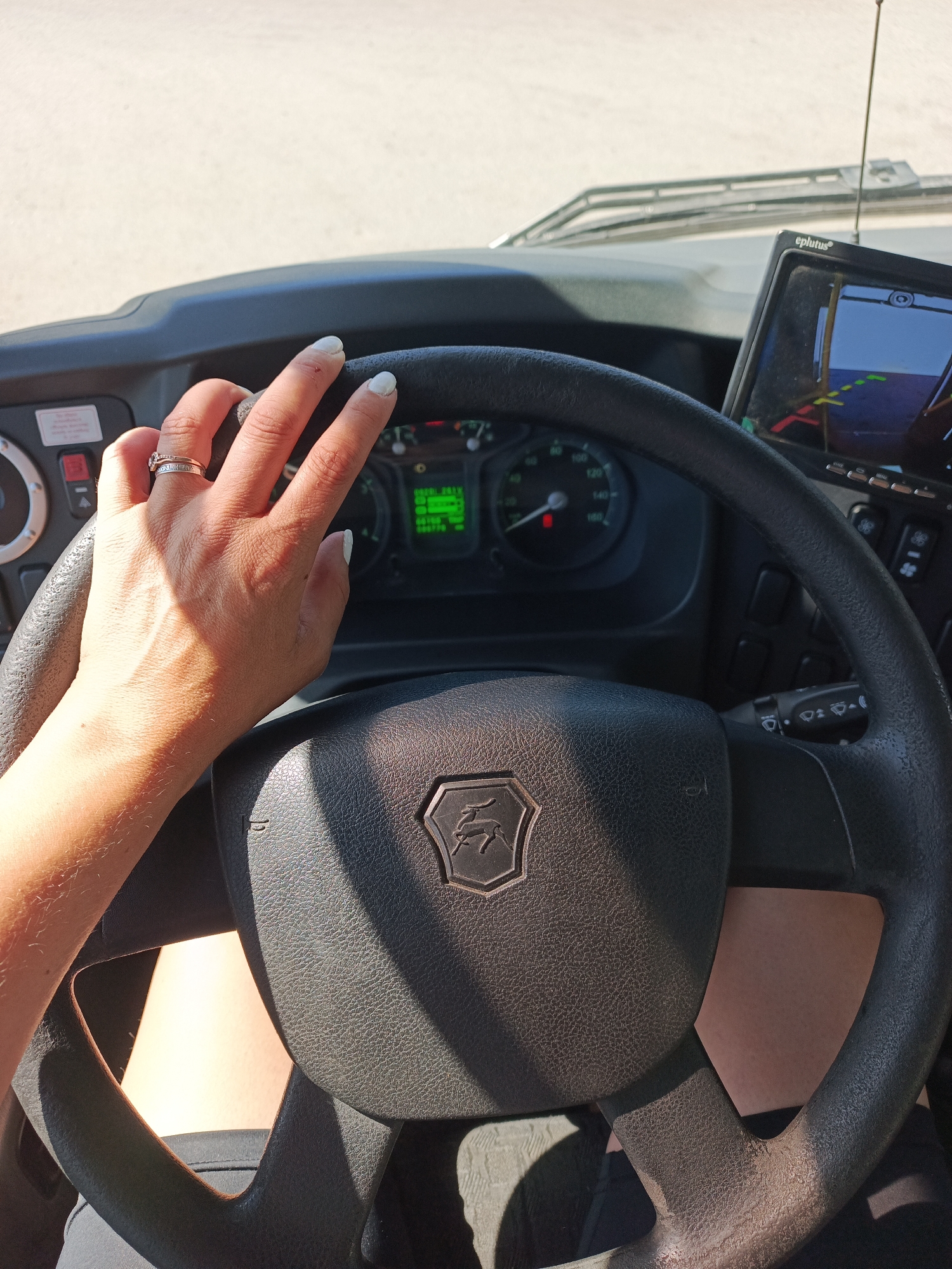 The image shows the interior of a vehicle, likely a bus or truck, with a focus on the driver's seat. The steering wheel, made of black plastic with a logo resembling a deer, is the central element. Behind the wheel, a dashboard with gauges and controls is visible. To the right of the steering wheel, there's a navigation system screen. A hand with a ring is resting on the steering wheel. The background is blurred, but the windshield and the outside environment suggest it's daytime. Based on the deer-like logo on the steering wheel, the vehicle is likely a GAZ (Gorky Automobile Plant) model. Given that GAZ vehicles are common in Russia, the location could plausibly be Chaikovsky, Russia, or any other Russian city. The person is operating the vehicle. The vehicle appears to be parked, based on the display on the navigation screen. The weather outside is clear and bright.
