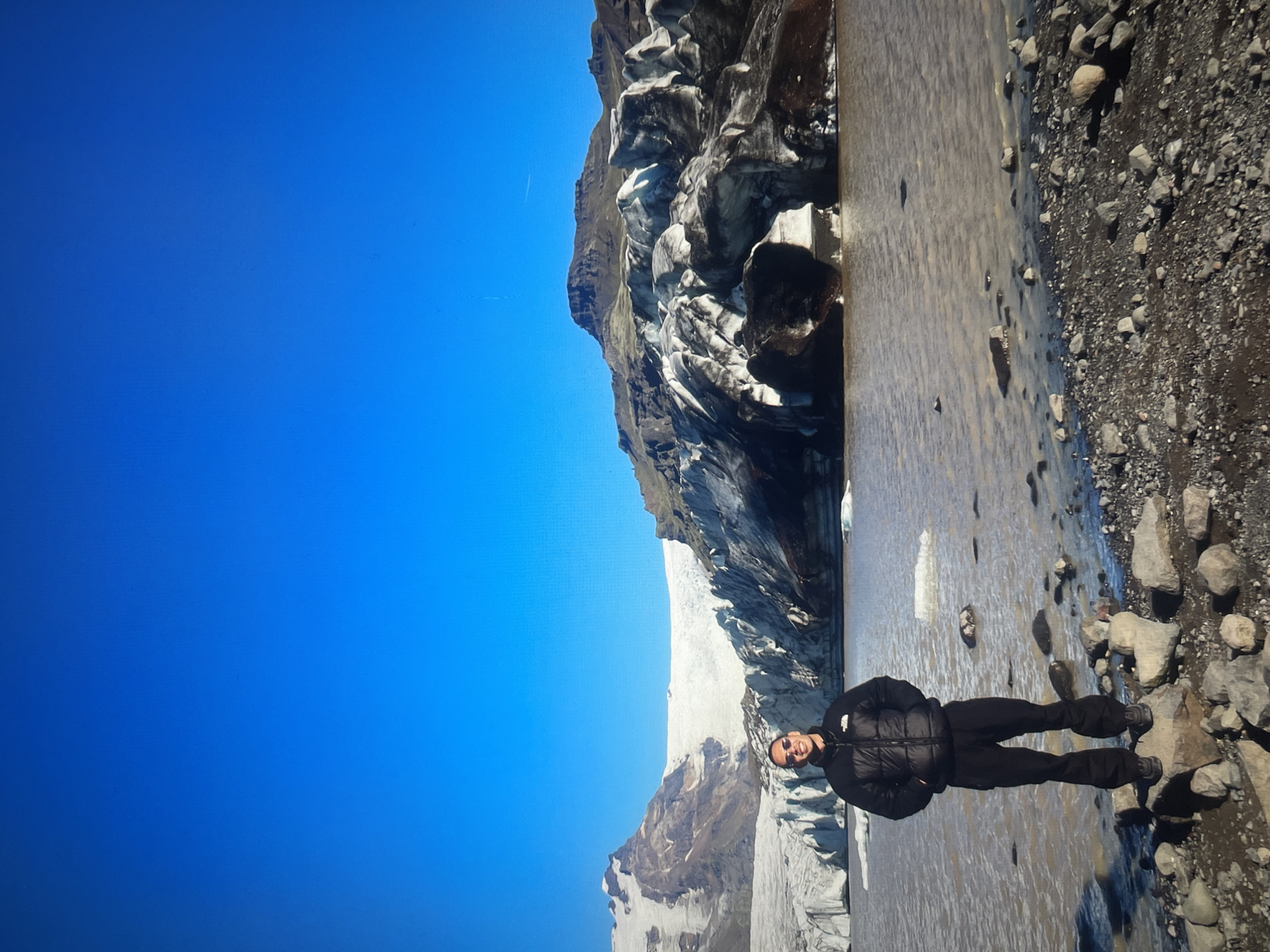 A photograph shows a man standing in a river next to a glacier. The man is wearing a black puffer jacket and black pants, and he is standing with his hands in his pockets. He is facing the camera and smiling. The river is murky and has a rocky bank. The glacier is large and white, with dark patches of rock and dirt. The sky is blue. The location is unknown.