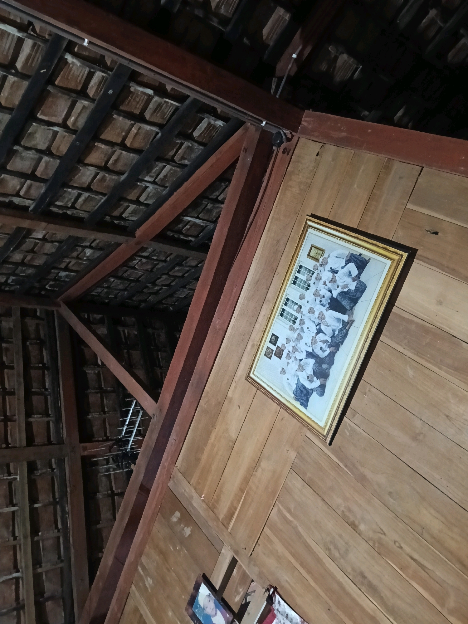 In Galuk, Indonesia, within a room constructed of wood, a framed photograph hangs on a paneled wooden wall. The photograph depicts a group of people, likely students, in white attire, seated in rows. The room's interior is characterized by exposed wooden beams and a tiled ceiling. The perspective of the image is angled upwards, capturing the architectural features of the space.