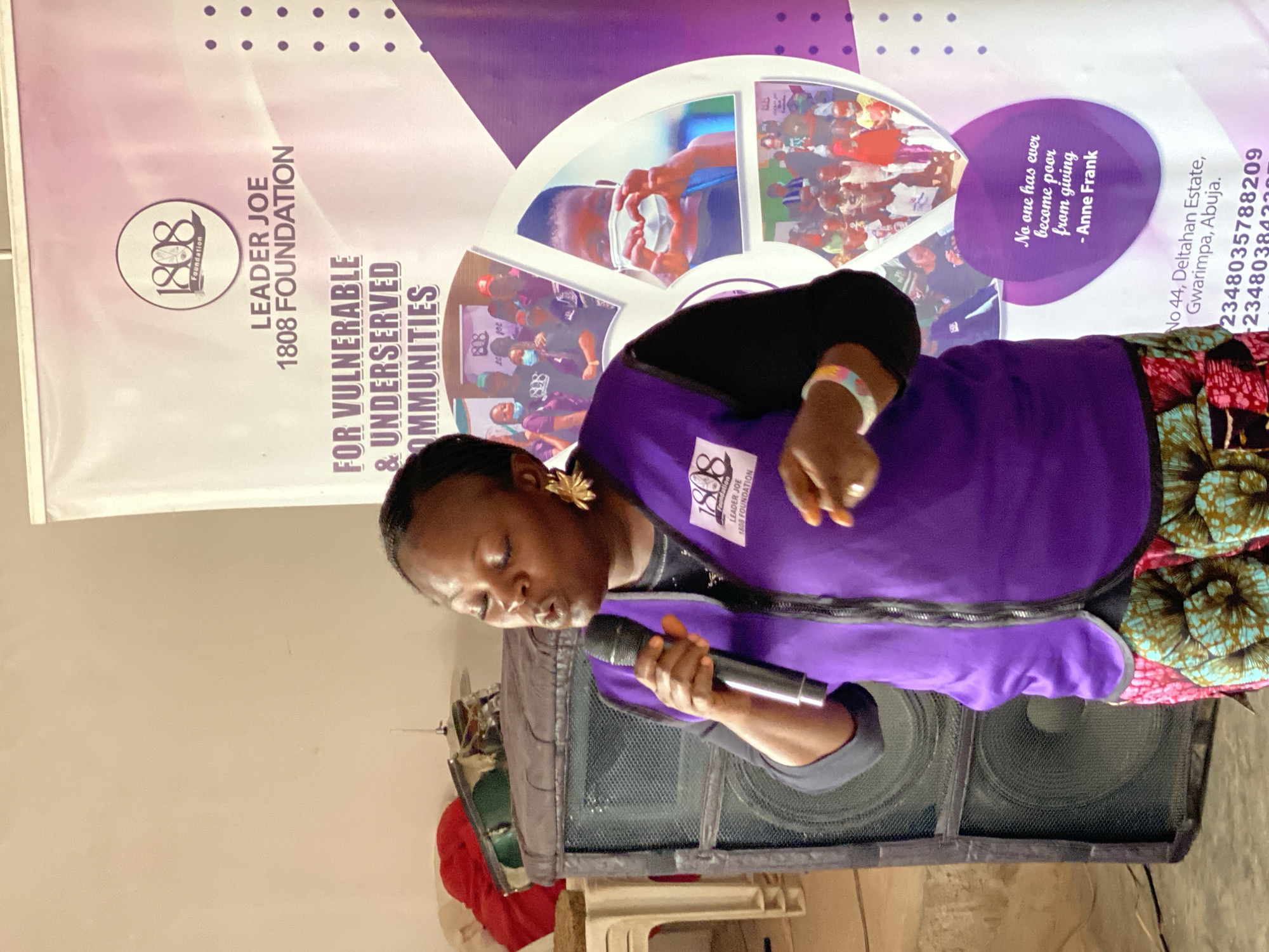 In a room with pale walls, a woman wearing a purple vest over a black shirt and a colorful patterned skirt is speaking into a microphone. She has her eyes closed and is holding the microphone in her right hand. The vest has a logo on the left chest. She is standing in front of a large black speaker system. To the right of the speaker is a large banner with the text "1808 FOUNDATION" and "LEADER JOE 1808 FOUNDATION FOR VULNERABLE & UNDERSERVED COMMUNITIES." The banner also features several circular images of people, including a child making a heart shape with their hands, and a quote by Anne Frank. Below the banner are the words "No 44, Deltahan Estate, Gwarimpa, Abuja." This indicates the location is likely within or associated with Gwarimpa, Abuja, Nigeria.