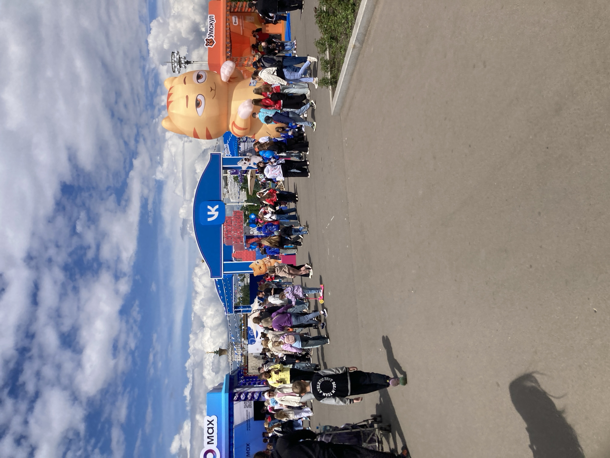 A street scene in Moscow, Russia, features people lining up under a series of promotional structures, including one shaped like a cat, under a bright cloudy sky. The structures feature the logos of VK, Umskool, and MAX. A section of paved ground extends from the right of the image. The foreground includes shadows cast by individuals standing outside the frame.