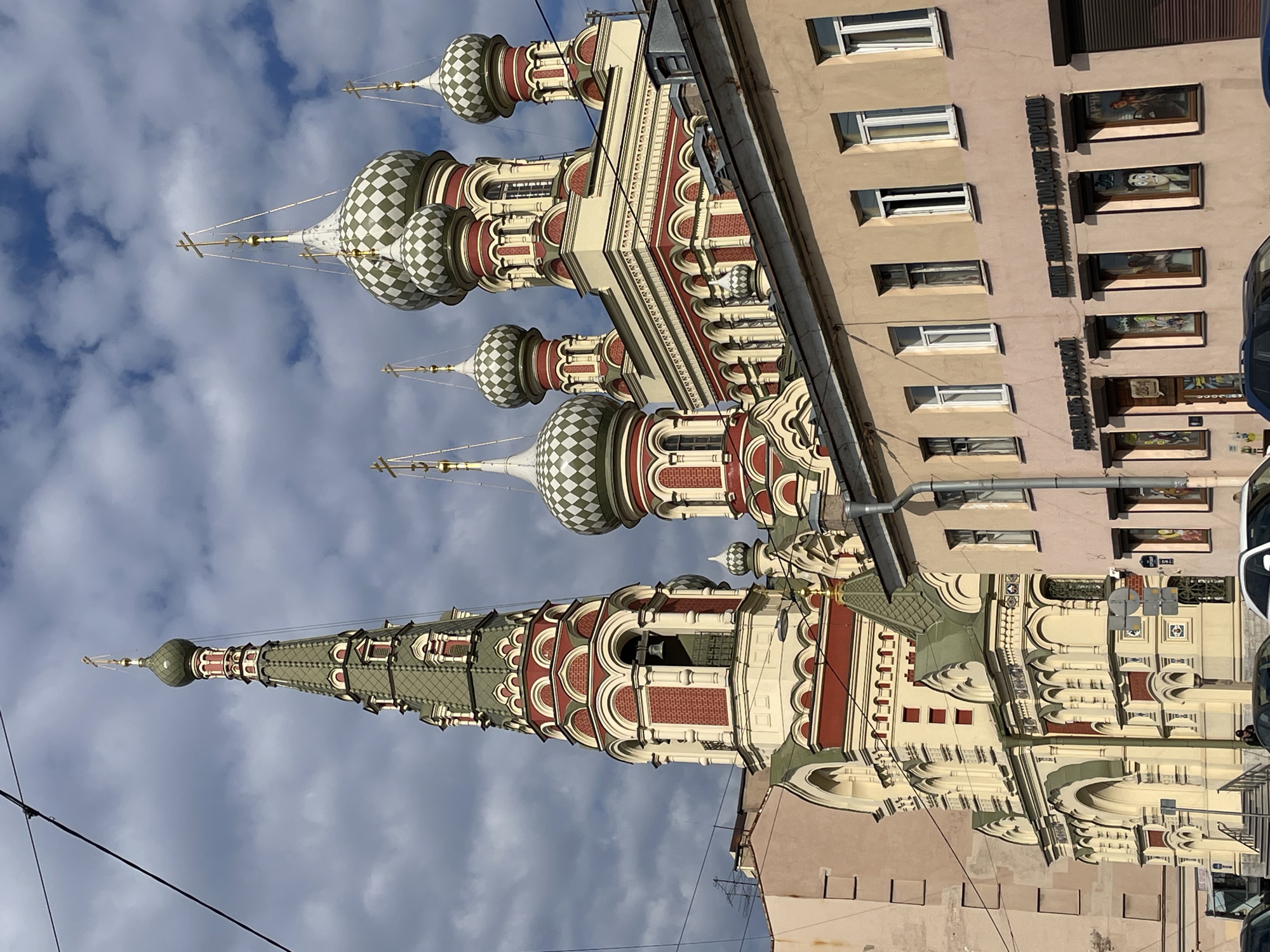 An eye-level shot captures a vibrant architectural scene in an urban setting, likely Saint Petersburg, Russia. The primary subject is an ornate building, characteristic of Russian Orthodox architecture, dominating the left side of the frame. It features multiple towers and onion domes, predominantly in cream, red, and green hues, with the domes adorned with a distinctive white and green checkerboard pattern. Golden crosses top the domes and spires, reaching towards a partly cloudy sky. The tallest spire boasts a green, scaled roof texture.
Adjacent to this grand structure, on the right, stands a multi-story building with a plainer, light brown facade and numerous rectangular windows. Along its side, various signs with Cyrillic text are visible. The most prominent signs read "АНИЧКОВ ДВОР" (Anichkov Dvor), "БАЛЕТНЫЕ КОРПУСА" (Ballet Buildings), and partially "УЧИЛИЩЕ РУССКОГО" (School of Russian...), suggesting a ballet school or related institution. Some ground-level windows display artwork or posters.
The sky above is bright with scattered clouds and patches of blue. Electrical wires crisscross the lower part of the frame, hinting at a street-level perspective. A small section of a white vehicle is visible on the far right. The overall impression is a sunny, daytime scene, highlighting the detailed craftsmanship of the older architecture against the backdrop of a functional urban environment. No specific activity or event is taking place; the image is a static portrayal of the buildings.