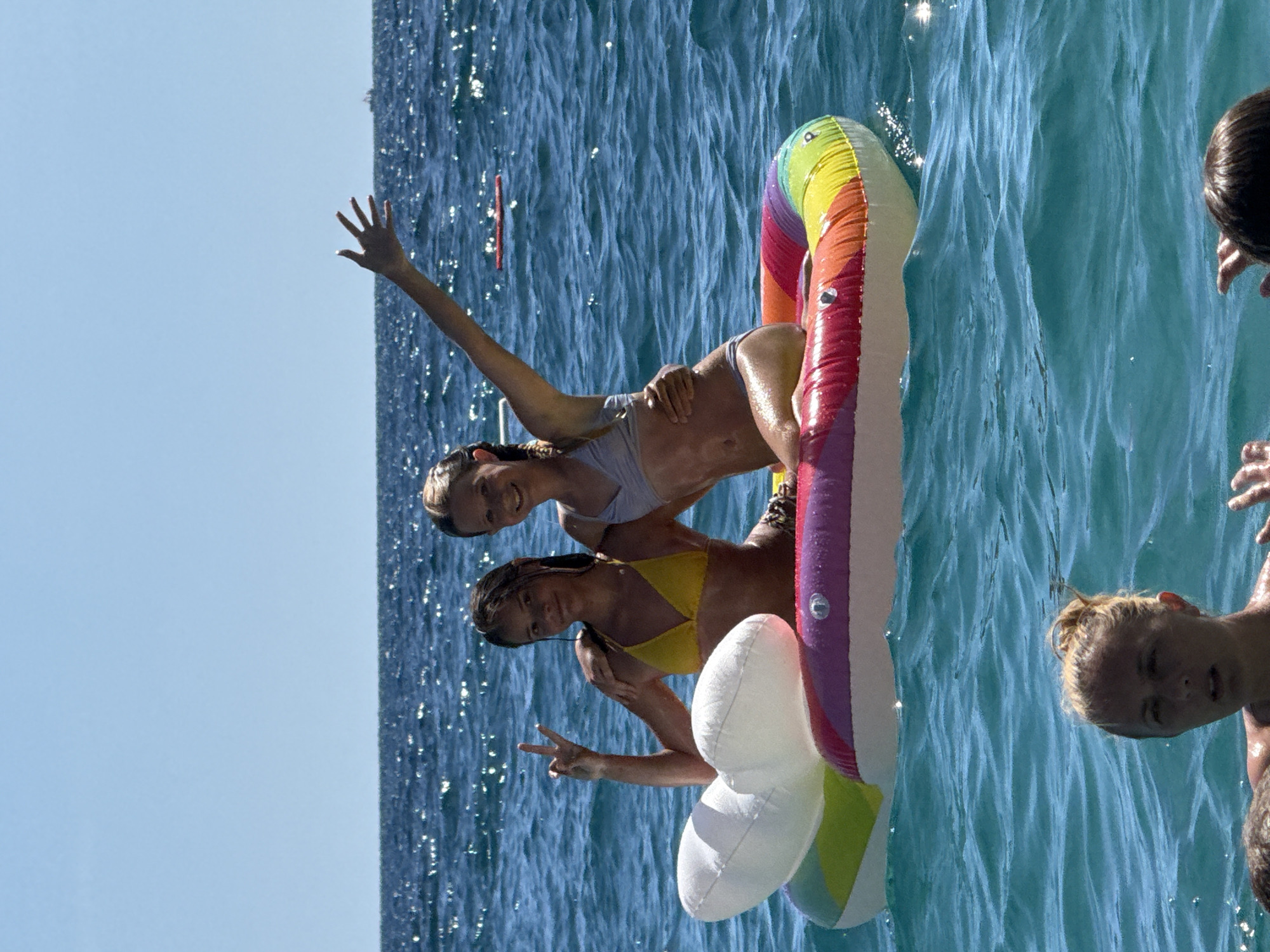 Two young women are pictured on a colorful inflatable in the Adriatic Sea off the coast of Kamenovo, Montenegro. The woman on the left wears a yellow bikini and holds up two fingers in a peace sign. The woman on the right wears a gray bikini and waves with her right arm. Both women are smiling. The sea is a vibrant turquoise blue with small waves. In the foreground, another person's head and shoulders are visible. In the background, a red object floats in the water, and a distant boat is visible on the horizon. The sky is clear and blue.
