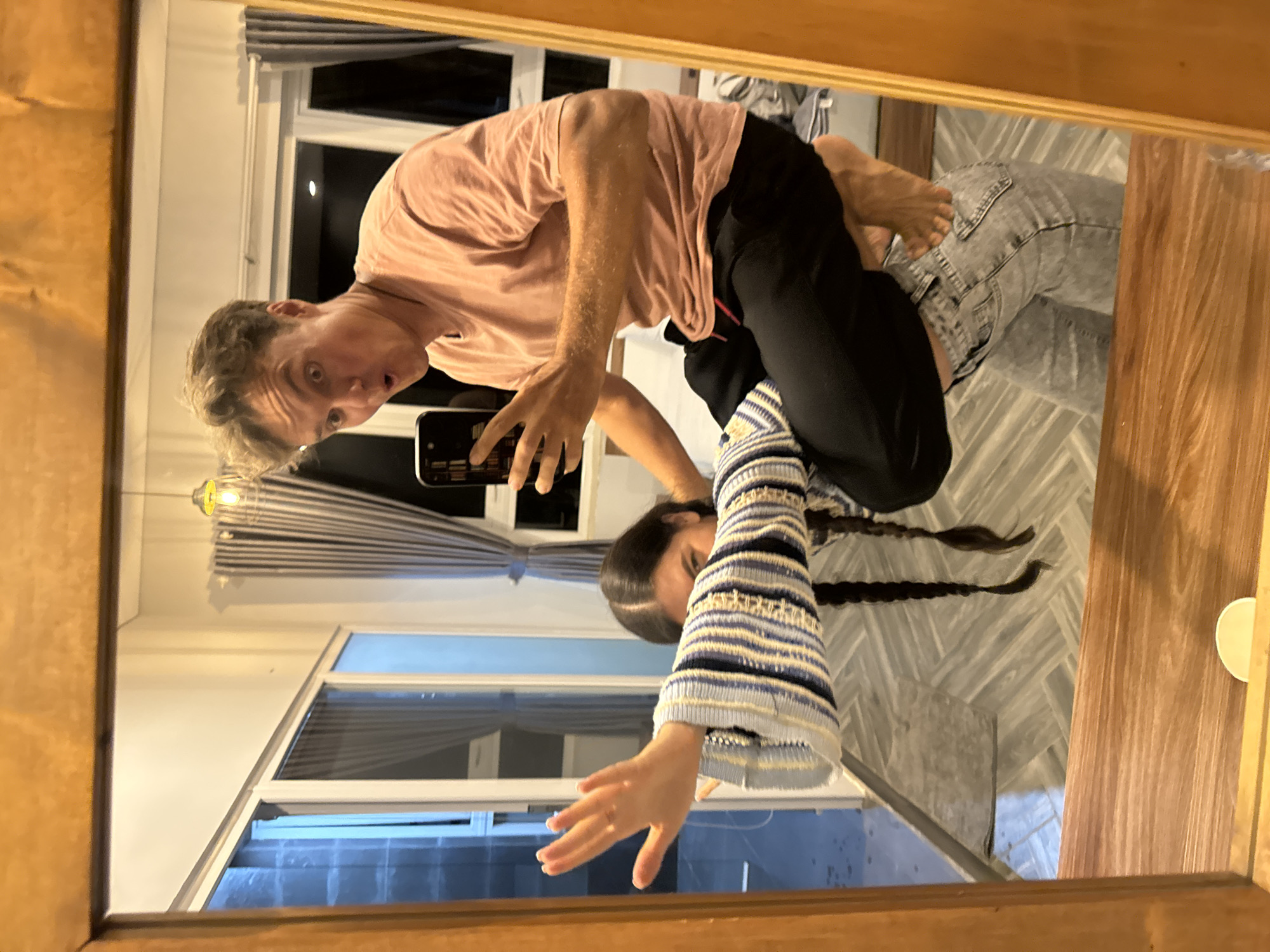 The image displays a mirror reflection of two individuals captured within a room. A man with short, light-colored hair and a wide-eyed, expressive face wears a light pink t-shirt and dark pants. He holds a black smartphone in his right hand, extended forward, seemingly taking a photo or video of their reflection.
Behind him, a woman with long, dark hair styled into two prominent braids is visible. She wears a striped long-sleeved top featuring patterns in blue, white, and yellow/beige. Her left arm is also extended forward in a similar pose to the man. Her lower body, partially obscured, appears to be wearing light grey jeans.
The mirrored scene reveals a room with light-colored walls and a patterned tiled floor in shades of grey. A large window with white frames shows a dark outdoor view, suggesting it is nighttime or evening, with a faint light source visible in the distance. Striped blinds are partially drawn over the window. A light fixture with a bulb hangs from the ceiling. The mirror itself has a warm-toned wooden frame, and a white cup is placed on the wooden surface to the right. The overall impression is one of a playful or spontaneous moment. The location is Da Lat, Vietnam. No discernible text is visible.