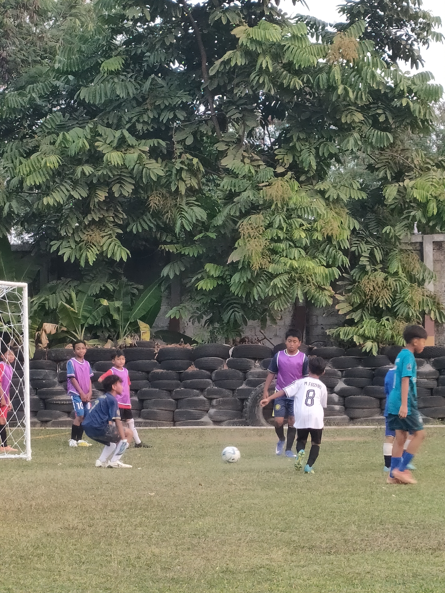 This image depicts a group of young boys playing soccer on a grassy field in Banguntapan, Indonesia. In the foreground, a boy in a white jersey with the number 8 and the name "M FAZIIO" is positioned, appearing to be either kicking or about to kick a soccer ball. He is wearing black pants and soccer cleats. To his left, another boy in a blue jersey and dark shorts is crouched low, possibly as a goalkeeper or preparing to make a save. Several other children, wearing brightly colored practice jerseys, are scattered across the field, engaged in the game. Behind the players, a wall constructed from stacked tires serves as a backdrop. Lush green trees with dense foliage dominate the upper portion of the image, with a white soccer goalpost visible on the far left. The scene suggests an informal soccer practice or game taking place outdoors.