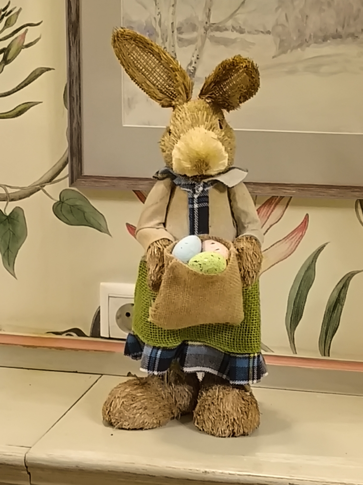 A decorative straw bunny figure stands on a surface, possibly a shelf or tabletop. The bunny is dressed in a light brown jacket over a blue plaid shirt, a green and blue plaid skirt, and holds a burlap bag filled with pastel-colored Easter eggs. The bunny has a straw head with large ears and a fluffy white nose. The background consists of a wall with floral wallpaper and a framed print of what appears to be a winter scene. A white electrical outlet is visible on the wall behind the figure. The scene is indoors, and the setting suggests a decorative or residential interior. The city of Smolensk, Russia is not evident from the scene.