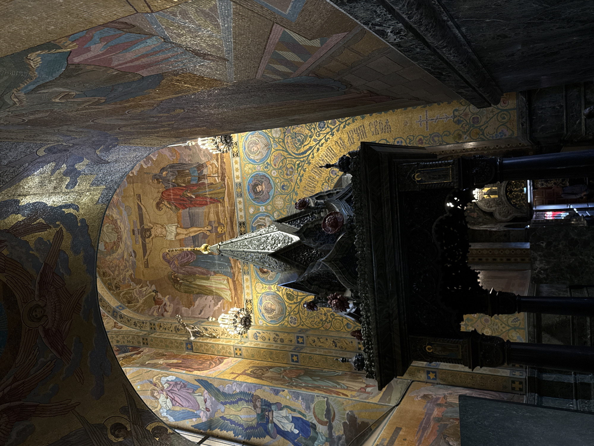 The image shows an ornate interior of a religious building in Saint Petersburg, Russia, likely a church or cathedral. The walls and ceiling are covered in intricate mosaics depicting religious scenes, including a crucifixion. A dark, elaborately carved structure, possibly an iconostasis or a tomb, is prominently displayed in the foreground. It is adorned with decorative elements and what appear to be floral arrangements. Crystal chandeliers hang from the ceiling, casting a warm light on the mosaics and architecture. The overall impression is one of grandeur and religious devotion.