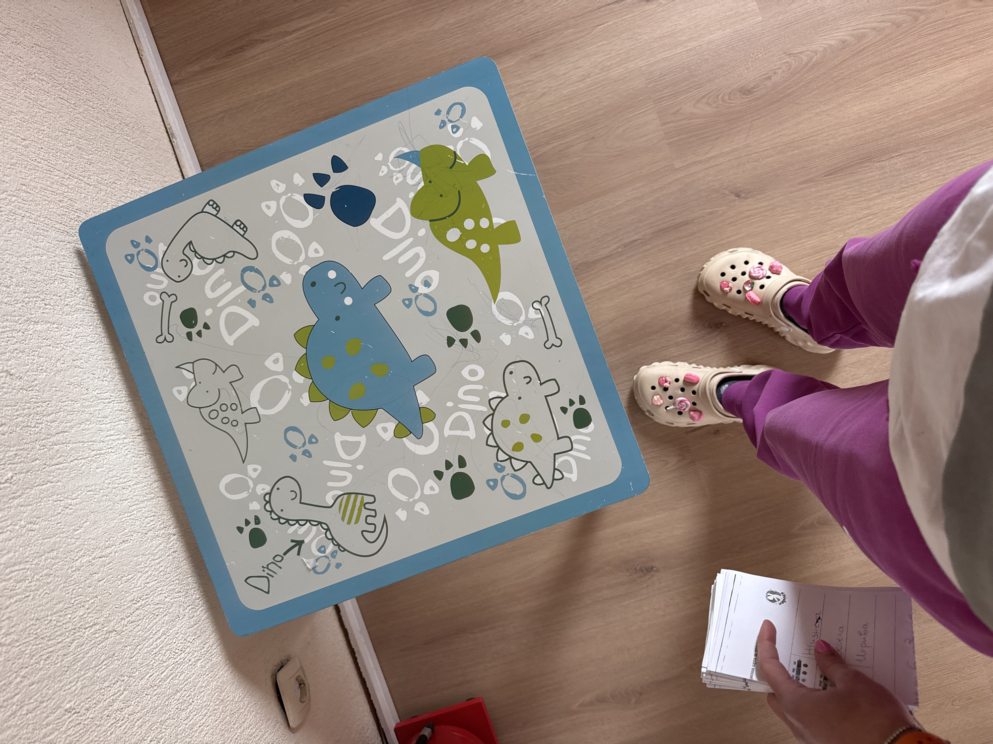 The scene depicts a person standing on a light-colored wooden floor next to a small, square children's table with a blue border and a dinosaur-themed design. The table is positioned near a wall with a textured, light-colored surface. The person is wearing purple pants and beige-colored shoes adorned with decorative charms. They are holding a stack of white cards with handwritten text in a non-English language. A red toy is partially visible under the table. There is no information provided to indicate this is in Sofia, Bulgaria.