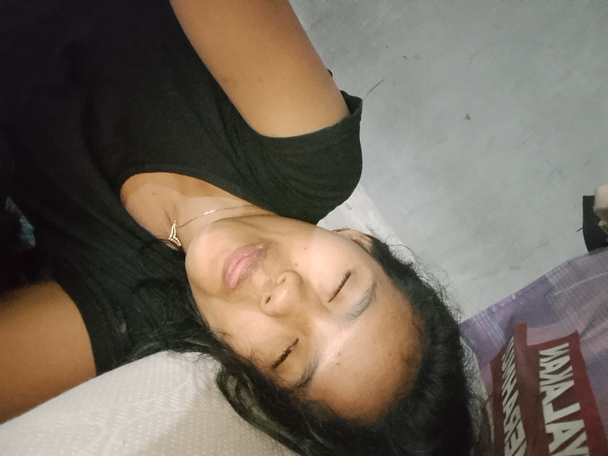 In Besuki, Indonesia, a young woman with dark hair lies on her side, her eyes closed. She is wearing a black t-shirt and a delicate necklace with a pendant. Her face is tilted slightly upwards, and her lips are parted, revealing a hint of her tongue. The background consists of a light gray, textured surface, possibly a wall or floor, and a portion of a patterned fabric, likely a blanket or sheet, in shades of purple and white. The lighting appears to be artificial, casting soft shadows. The overall impression is one of rest or sleep.