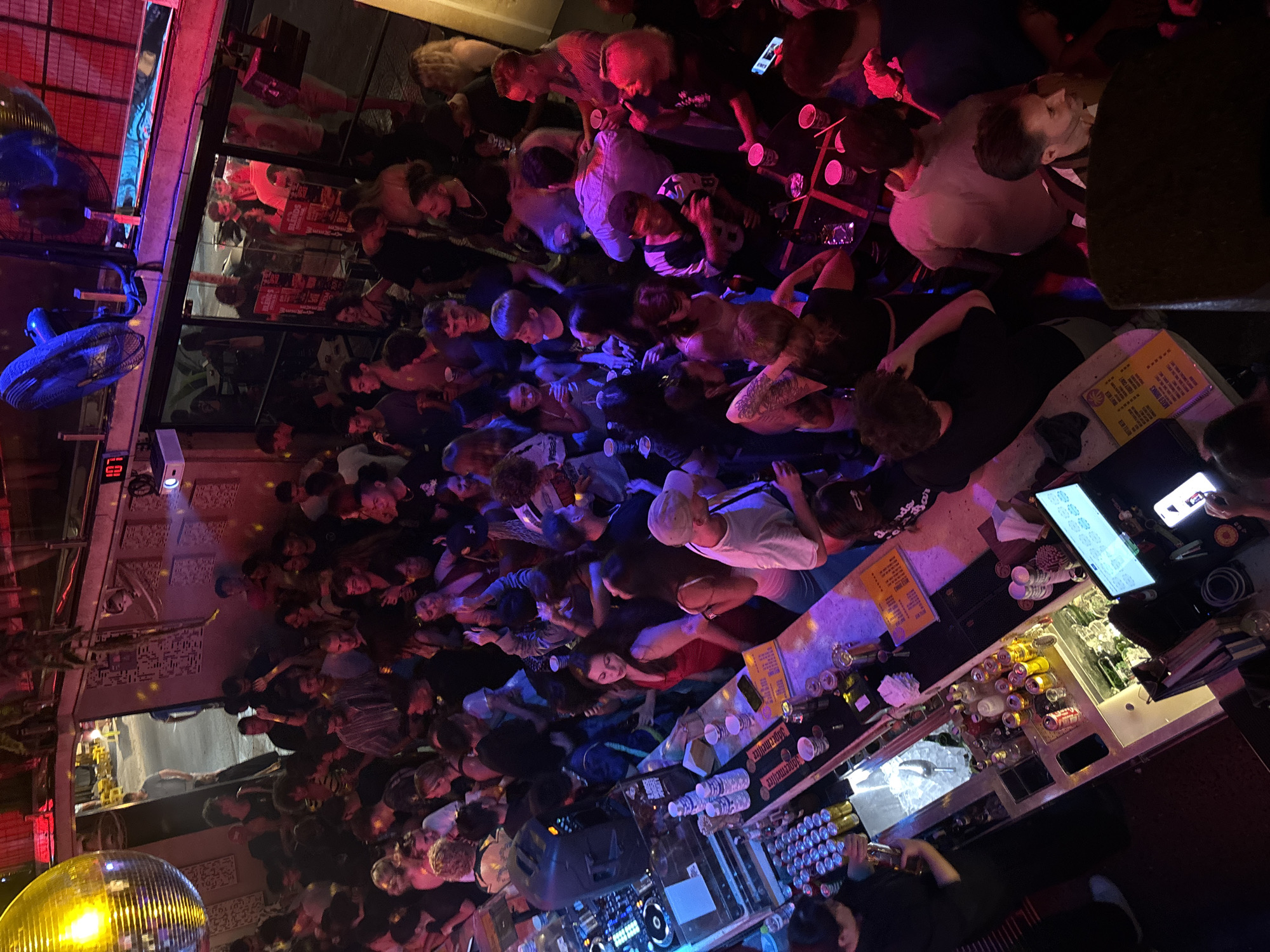 This image depicts a crowded indoor nightclub scene. The lighting is dim and colorful, with neon lights and a disco ball illuminating the space. The people present are engaged in various activities such as dancing, socializing, and ordering drinks from the bar.
The bar area is well-stocked with bottles of alcohol and cans of beverages. There are also several people working behind the bar, preparing drinks. The overall atmosphere appears lively and energetic. The presence of the number "107" displayed on a digital clock suggests this might be a specific venue or a room within a larger establishment.
Given the prompt's request for location in Da Nang, Vietnam, it is possible this establishment is a nightclub or bar within that city. Without further context or visual cues specific to Da Nang, the exact location cannot be definitively determined.