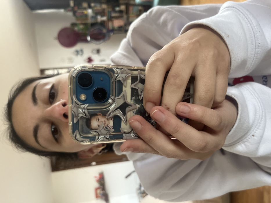 A person holds an iPhone with a clear case. The phone case has a picture of a young child surrounded by silver stars. The person wears a white sweatshirt. A blurred background shows a room with miscellaneous objects. The individual is holding the phone up to take a selfie. The phone is angled slightly, showing the camera lenses. The person's face is visible, looking directly at the camera.