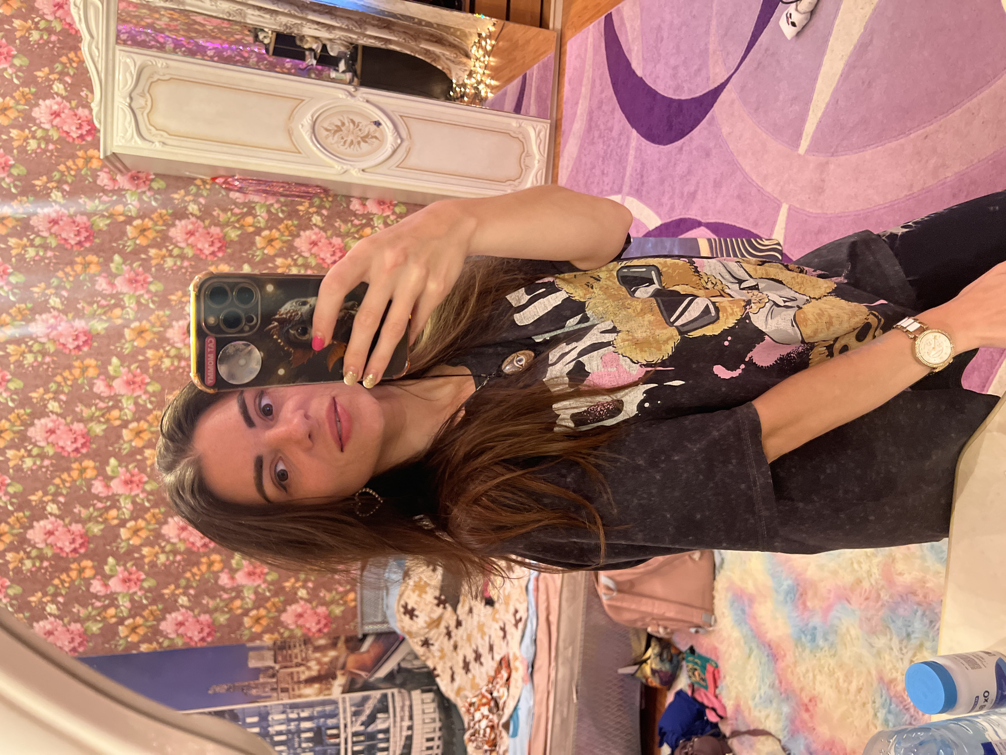 The media file is a portrait-oriented, slightly tilted, vertical selfie taken in a bedroom. The primary subject is a young woman with long, brown hair and fair skin, holding a smartphone to take the picture. She is wearing a dark grey t-shirt with a graphic print featuring cartoonish figures in sunglasses and a gold-colored watch on her left wrist. Her fingernails are painted in vibrant pink and gold glitter.
The background of the room is busy and eclectic. To the left, there is a wall covered in floral wallpaper with pink and orange flowers on a brown background. Behind the woman and to the right, a large, ornate white furniture piece, possibly a wardrobe or vanity, is visible, with decorative moldings and an oval inset. The reflection in a mirror above this furniture shows part of the room, including what appear to be string lights. A purple rug with a swirling, abstract design covers a portion of the floor. A light pink backpack rests on the floor near a heap of colorful clothing. Further into the background, a fuzzy rug with pastel colors of pink, blue, and yellow is visible, along with what seems to be a building mural or poster.
The lighting suggests it is daytime, possibly indoors with artificial lighting contributing to the overall brightness. The woman's expression is neutral, with her eyes looking directly at the camera. The overall impression is a candid moment of self-documentation within a personalized and somewhat maximalist living space. The location context is Krasnoe-na-Volge, Russia.