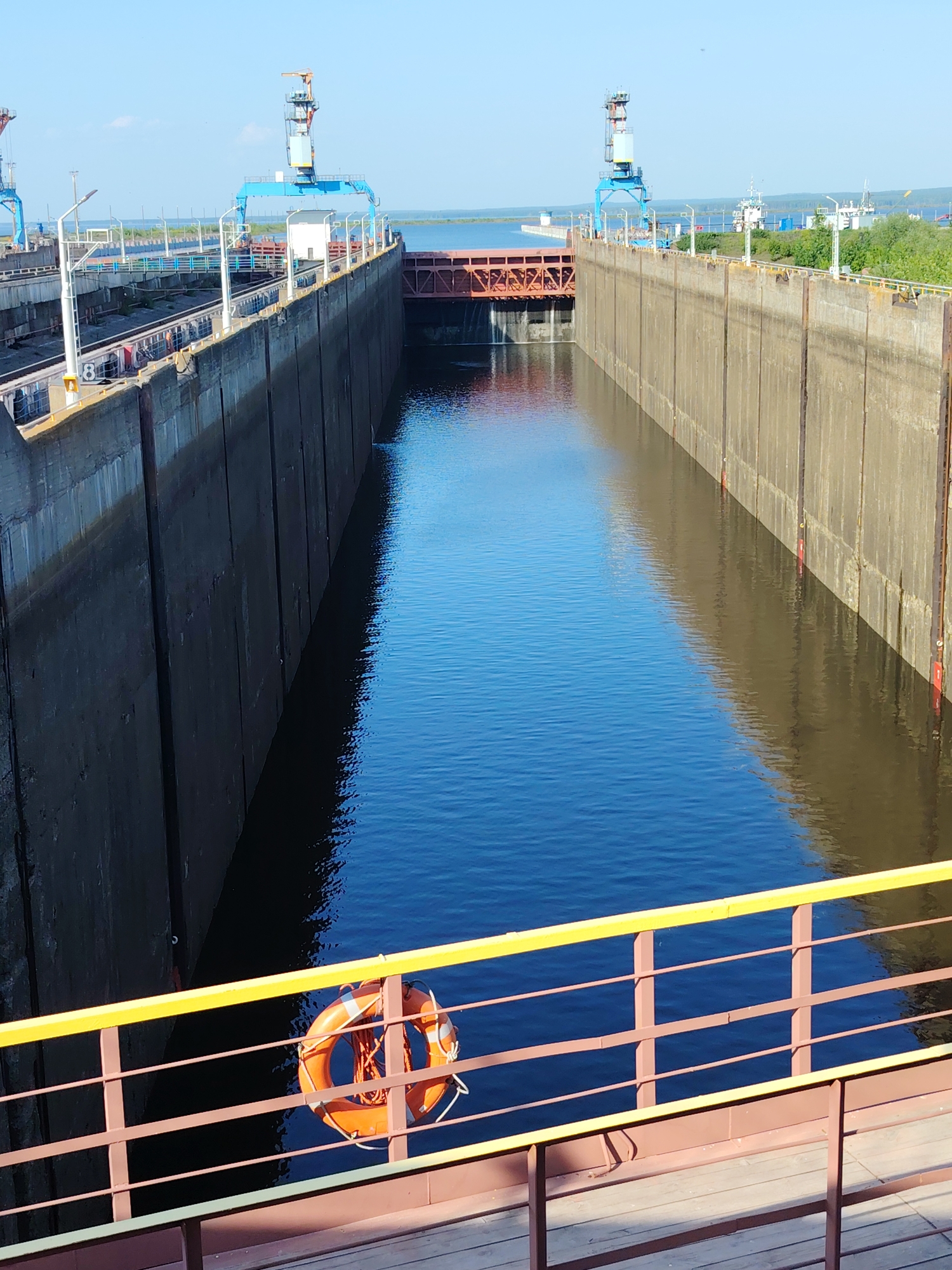 The image shows a section of the Kama River lock, located in the city of Chaikovsky, Perm Krai, Russia. The image looks towards the end of a canal created between two concrete lock walls filled with still, dark water. The walls stretch along the canal, with two blue-painted gantry cranes visible beyond the end of the lock. At the end of the lock is a section of brick or metal structure across the water. In the foreground, an orange life preserver hangs on a brown metal railing with a yellow top bar. The scene appears to be taken on a sunny day.