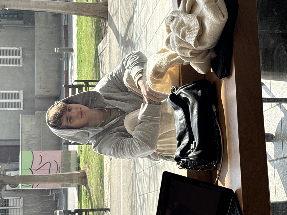 The image depicts a young male seated indoors at a wooden counter, looking out through a large window to a street scene in Tel-Aviv, Israel. The male, appearing to be in his late teens or early twenties with light brown hair, is wearing a light grey hooded sweatshirt over an olive-green shirt and light beige pants. He is seated with his knees bent, resting his right hand near his left knee, and has a ring on his right ring finger. He looks directly towards the viewer with a neutral to slightly smiling expression.
On the wooden counter in the foreground, from left to right, are: an open black laptop with a dark screen, a black leather handbag with a metallic chain strap, and a loosely folded light-colored (beige/cream) knitted sweater or blanket resting on a small black object. A white cable is visible extending near the handbag.
Through the window, a paved sidewalk, a strip of green grass, and two deciduous trees with bare branches are visible. In the background, a light grey building with two windows fitted with white louvered shutters is present. To the left of the building, a light green utility box or cabinet with a pink abstract drawing is visible. Dark-colored street furniture, including benches and bollards, can also be observed. The scene is illuminated by natural daylight.