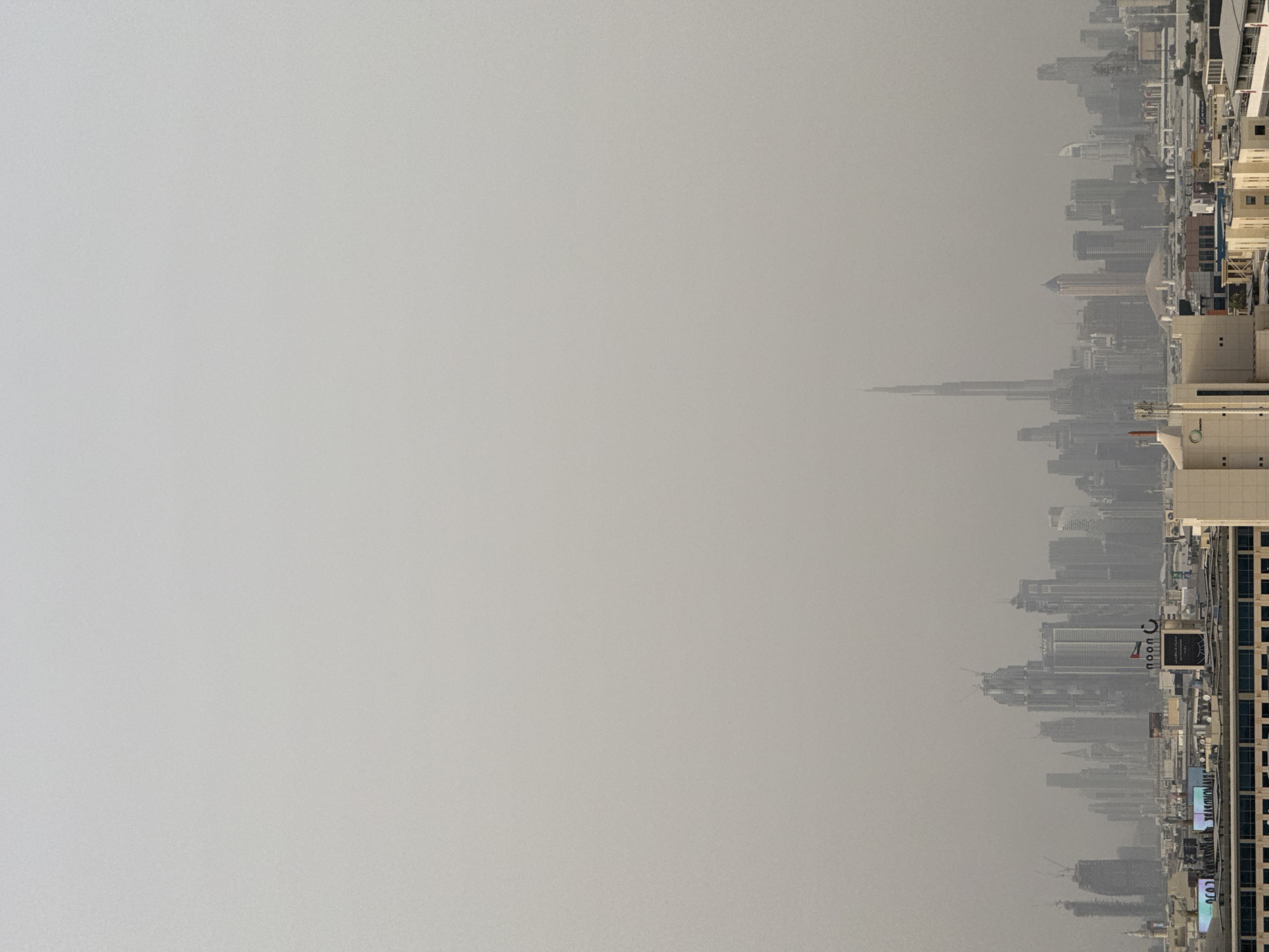 The image shows the city of Dubai, United Arab Emirates on a hazy day. The Burj Khalifa is visible in the skyline. Other skyscrapers and buildings are also present. The sky is a uniform grey color due to the haze.