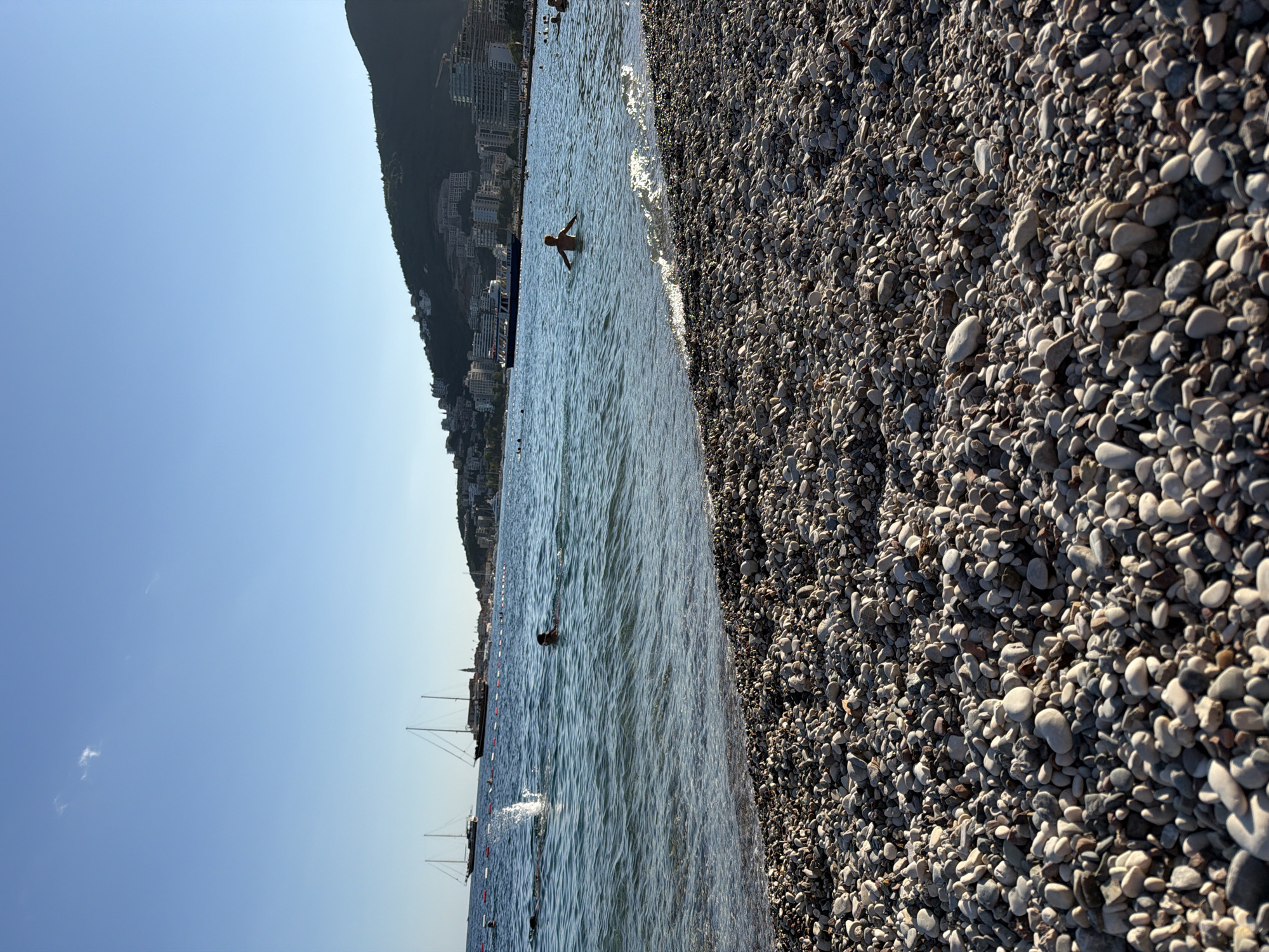 This is a photograph taken from a pebble beach in Budva, Montenegro. The foreground is dominated by the textured surface of the beach, composed of numerous light-colored, smooth pebbles. In the middle ground, the clear blue water of the Adriatic Sea laps gently at the shore. Several people are visible in the water; one individual is swimming, and another is standing with their arms outstretched. In the background, a line of boats, including sailboats and a ferry, are anchored or moving on the sea. Beyond the water, the cityscape of Budva is visible, with buildings cascading down the hillside of a lush green mountain. The sky is a clear, bright blue, with a few wispy clouds. The overall scene depicts a serene summer day at a popular coastal resort.