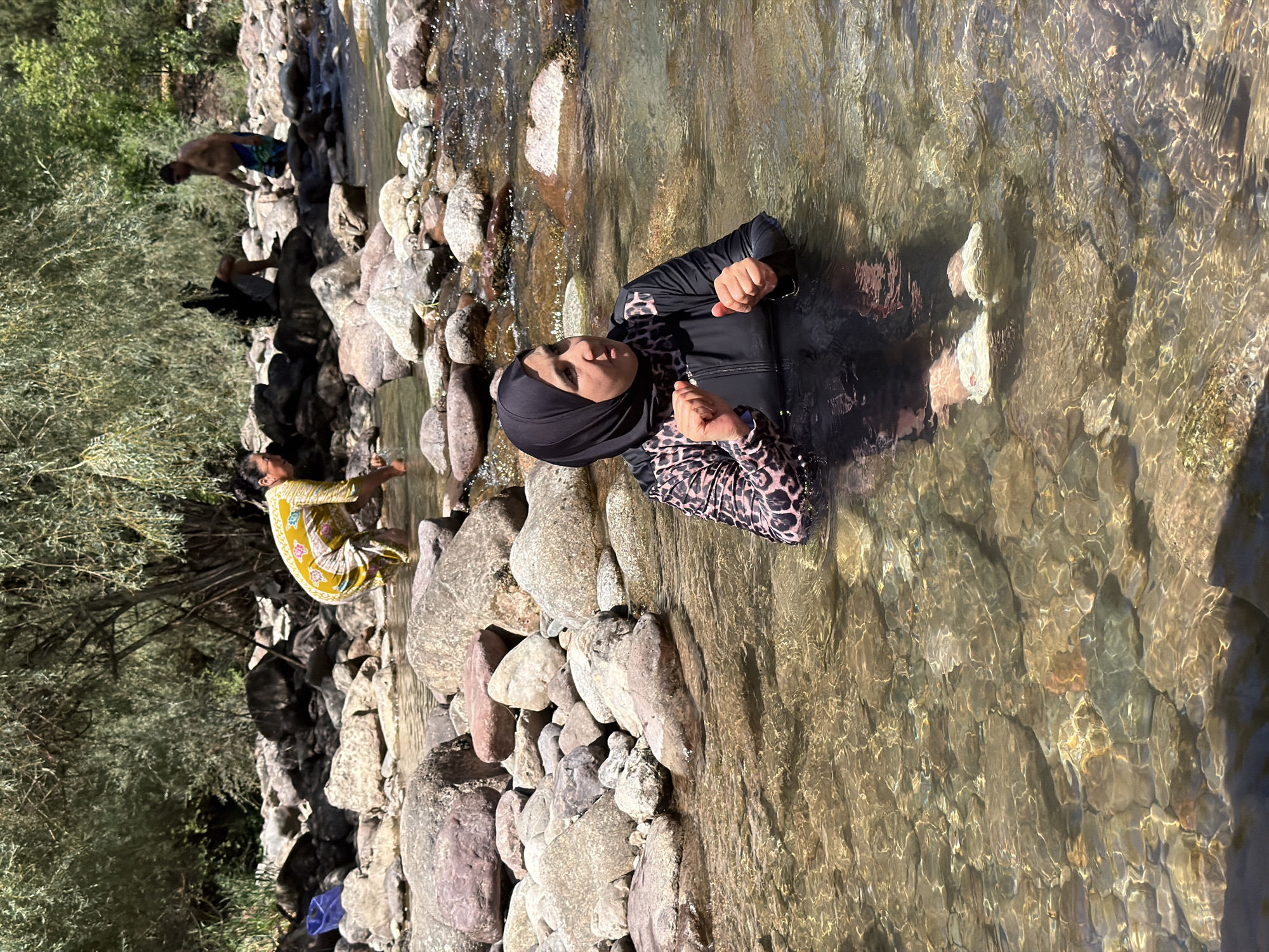 In the city of Khumsan, Uzbekistan, a woman wearing a black hijab and a leopard-print swimsuit is submerged in a clear, rocky river. She is facing to the right, with her hands clasped in front of her. Her feet, clad in white water shoes, are visible beneath the surface of the water. Behind her, another person in a yellow and white patterned garment is washing their hands in the river, while further in the background, two men are standing by the riverbank. The environment is characterized by large, smooth rocks lining the riverbed and a backdrop of lush green trees.
